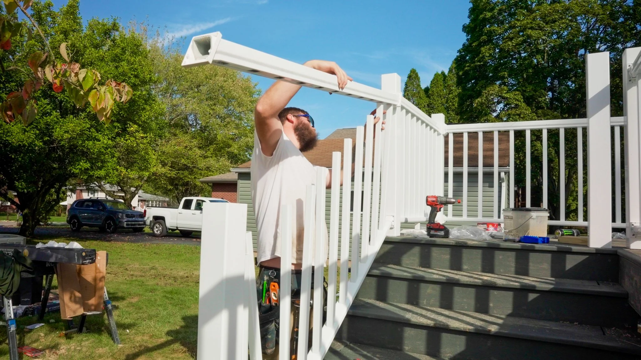 A man installing a white staircase at a residential construction site outdoors, with trees, a house, and cars visible in the background