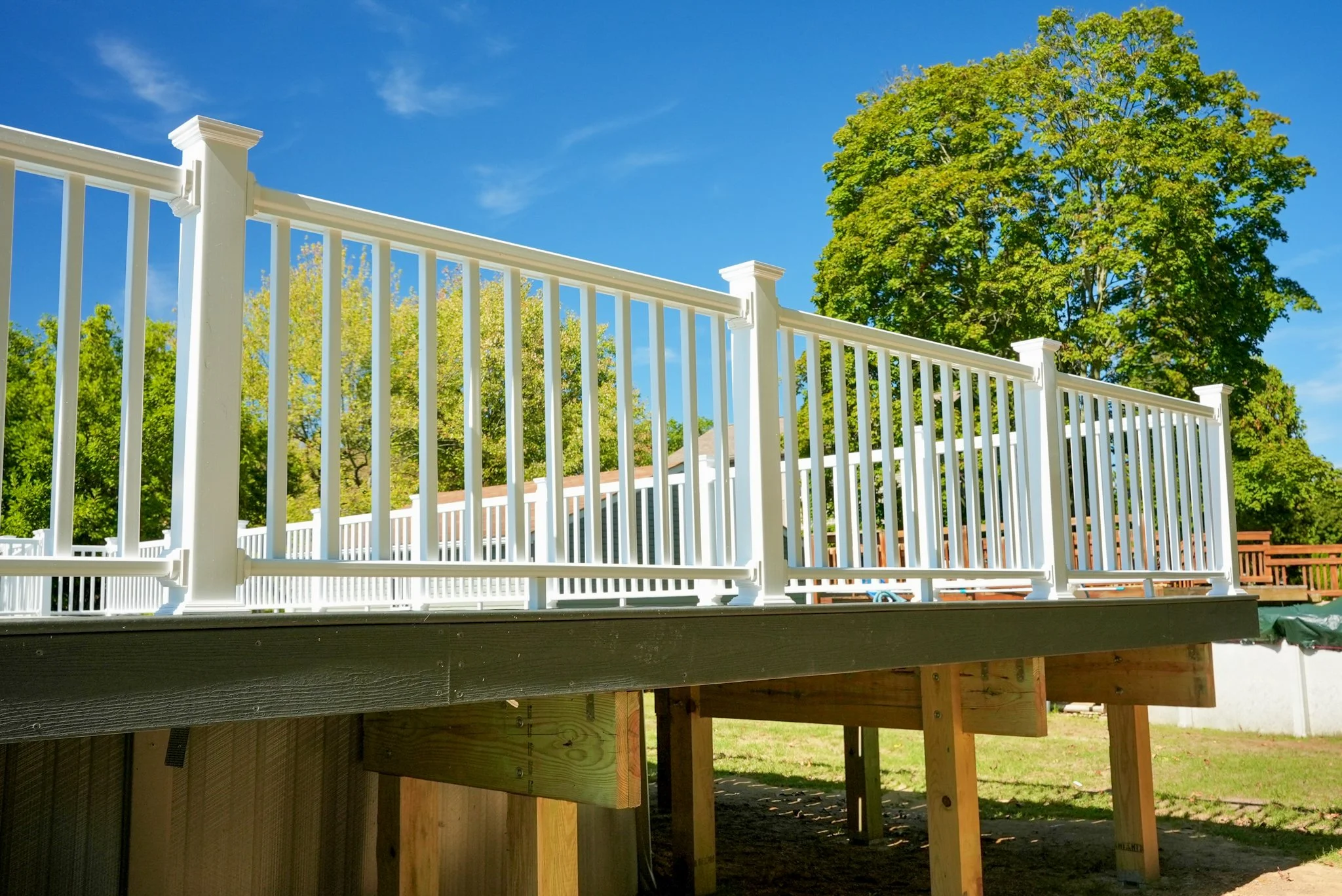 New white fence surrounding a deck under construction, with trees and blue sky in the background.