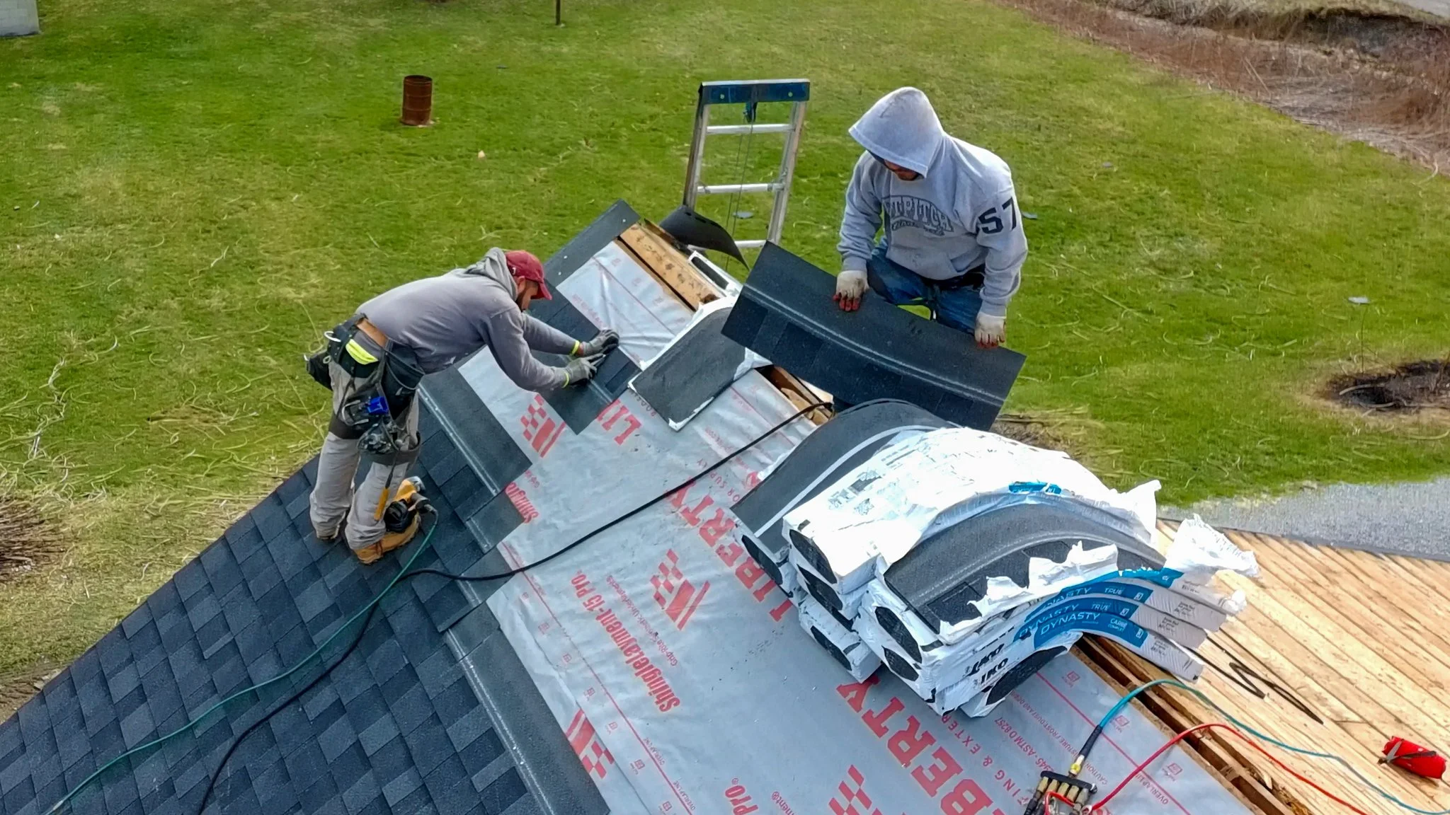 Two Pittsburgh roofers working outdoors, one with a white hoodie with a business logo and the other with a red hoodie, working on a roof repair project.