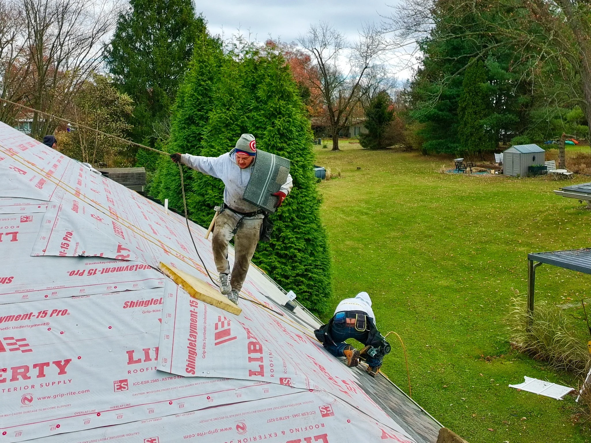 Two Pittsburgh workers installing roofing shingles on a sloped roof, one standing and holding a rope, the other kneeling and using tools, with a green backyard and trees in the background.