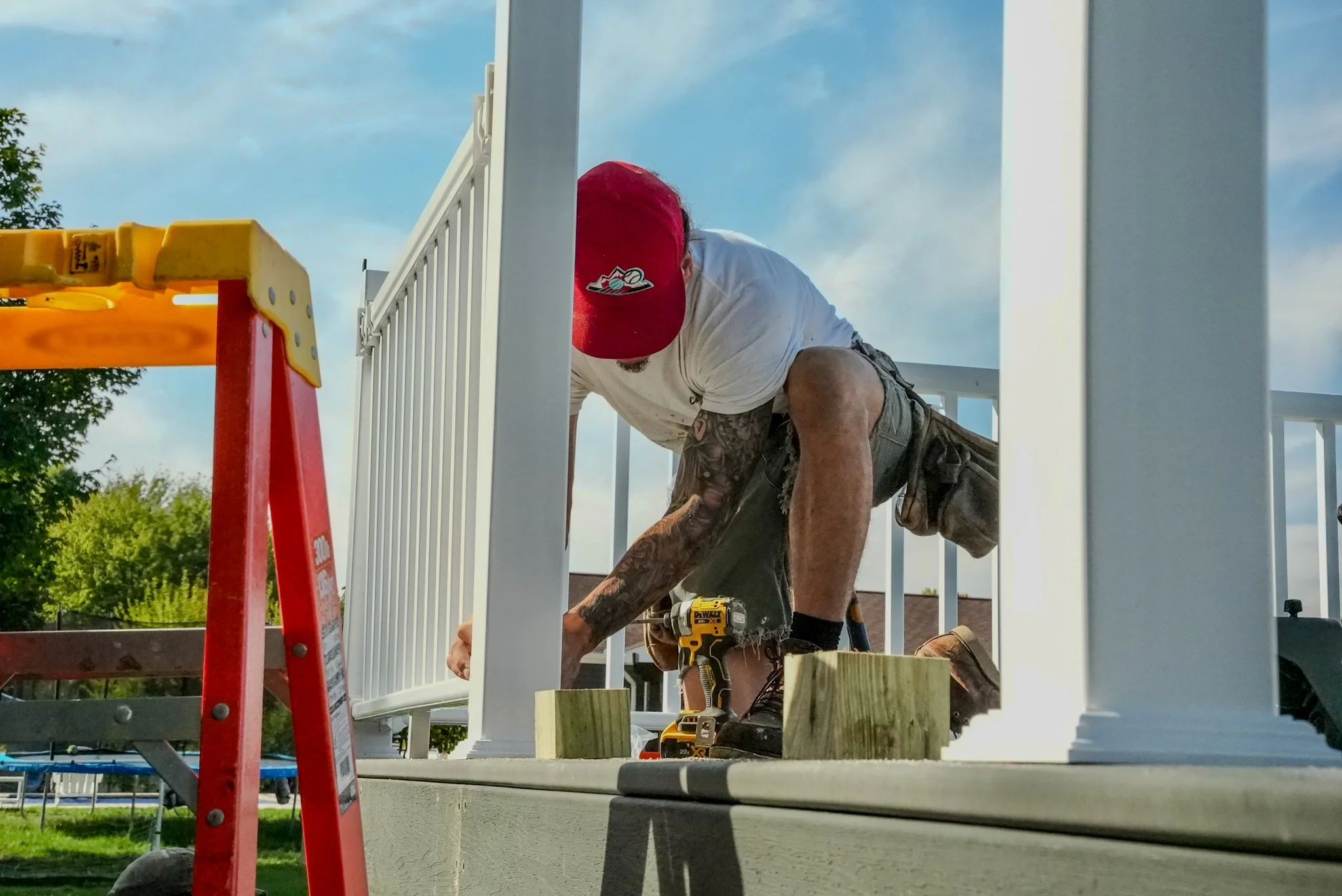 A man installing a white balcony railing outdoors, kneeling on a platform with tools and wooden blocks, wearing a red cap, white shirt, and shorts.