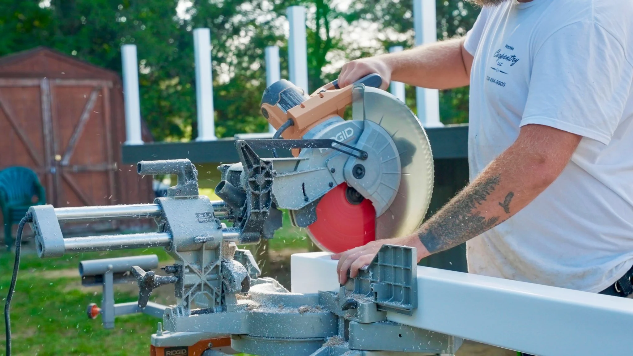 A man with tattoos on his arm using a sliding compound miter saw to cut a piece of wood outdoors, with sawdust flying and a wooden shed and trees in the background.