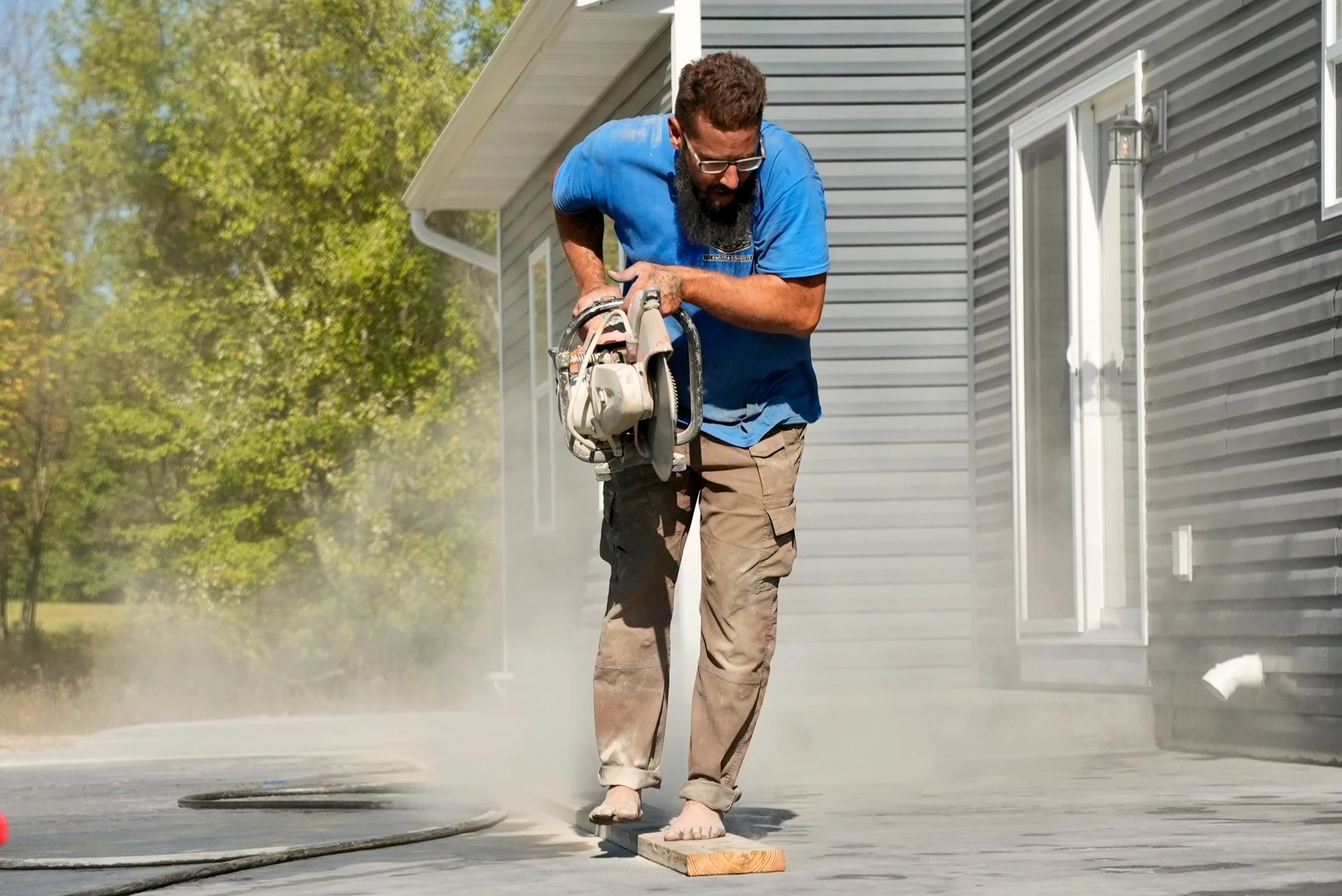 Pittsburgh contractor with a blue t-shirt, working with a power saw on a construction site outside a house with gray siding. Dust or sawdust is visible.