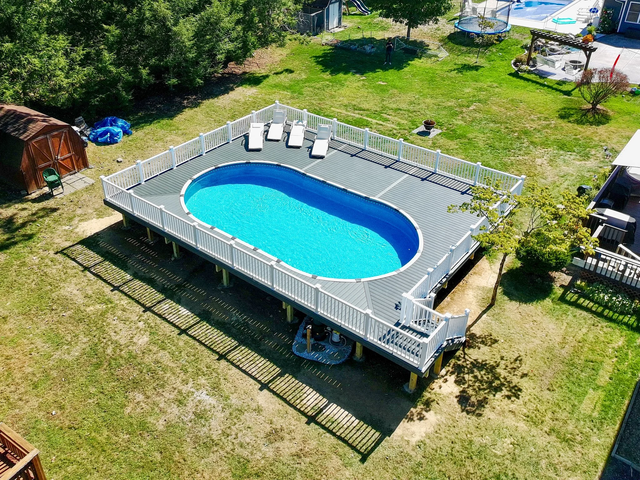 A backyard with an above-ground oval swimming pool on a raised deck with a white railing, surrounded by grass and trees, with lounge chairs on the deck and various outdoor furniture and toys in the yard.