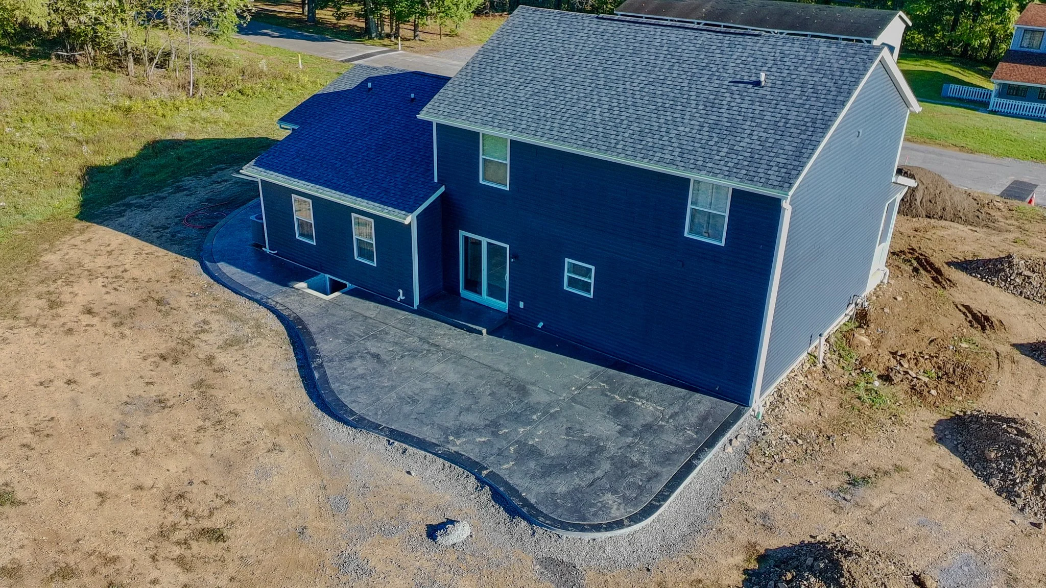 A newly constructed two-story blue house with a gray shingle roof, surrounded by an unfinished yard, with a concrete driveway in the front and some surrounding dirt areas, under a clear sky.