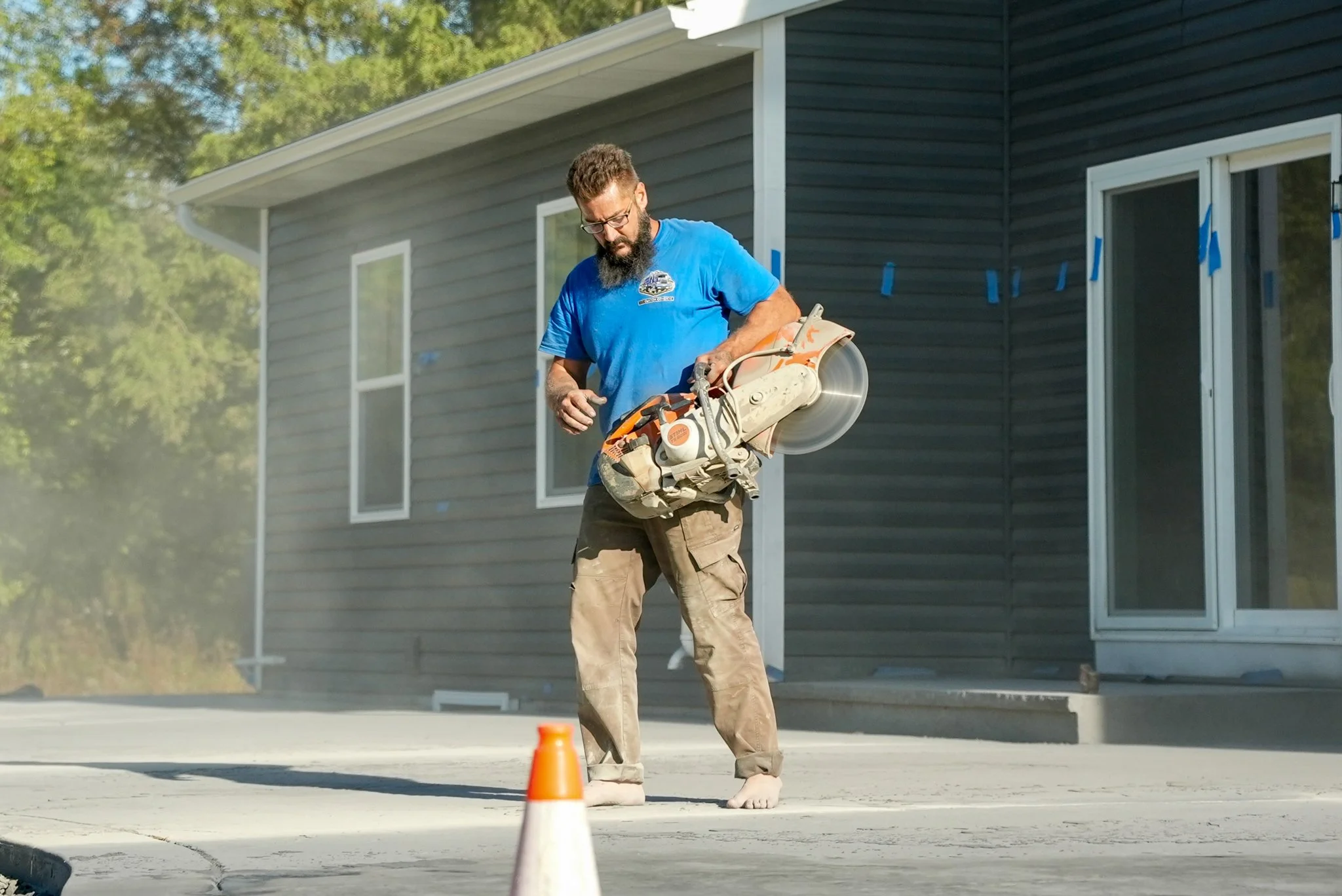 Pittsburgh contractor with glasses and beard using a concrete saw on a driveway outside a house, with a traffic cone nearby.