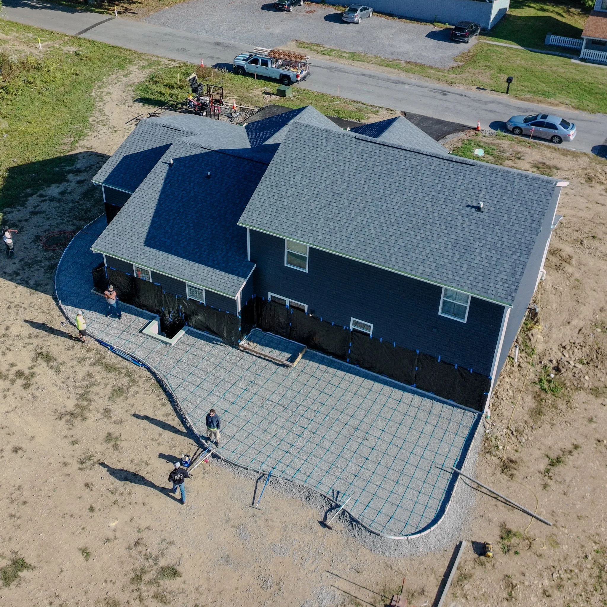Aerial view of a Pittsburgh house under construction with a new concrete patio and workers on site. The house has a gray shingle roof and dark siding, with construction materials and vehicles nearby.