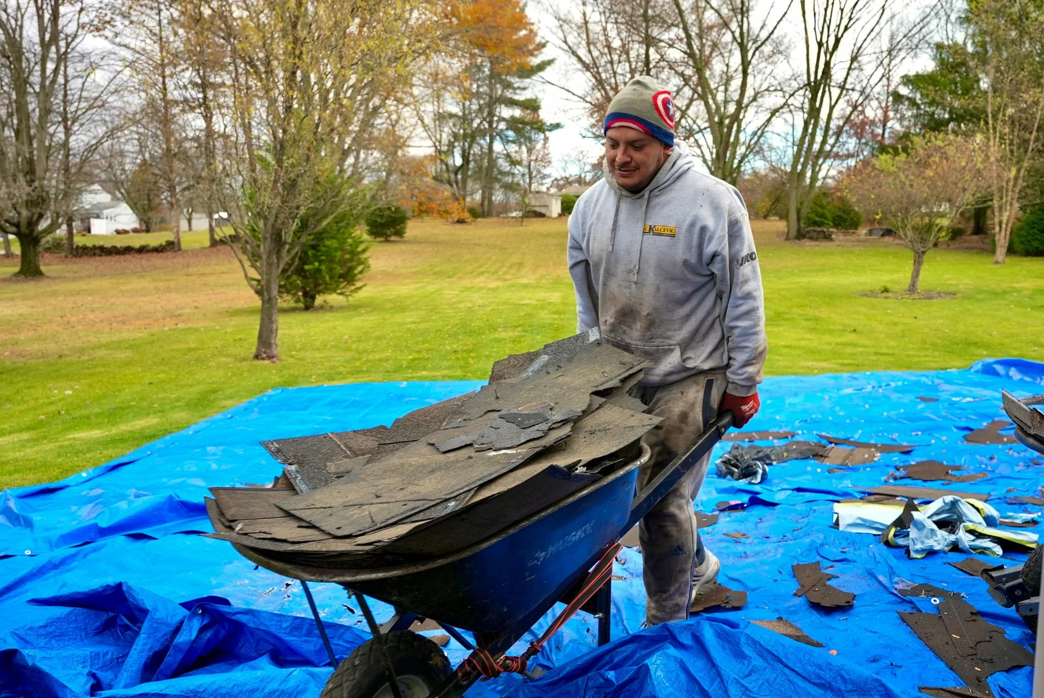 A Pittsburgh worker with a beanie and gray hoodie pushing a wheelbarrow filled with broken asphalt tiles outdoors on a blue tarp in a grassy backyard.