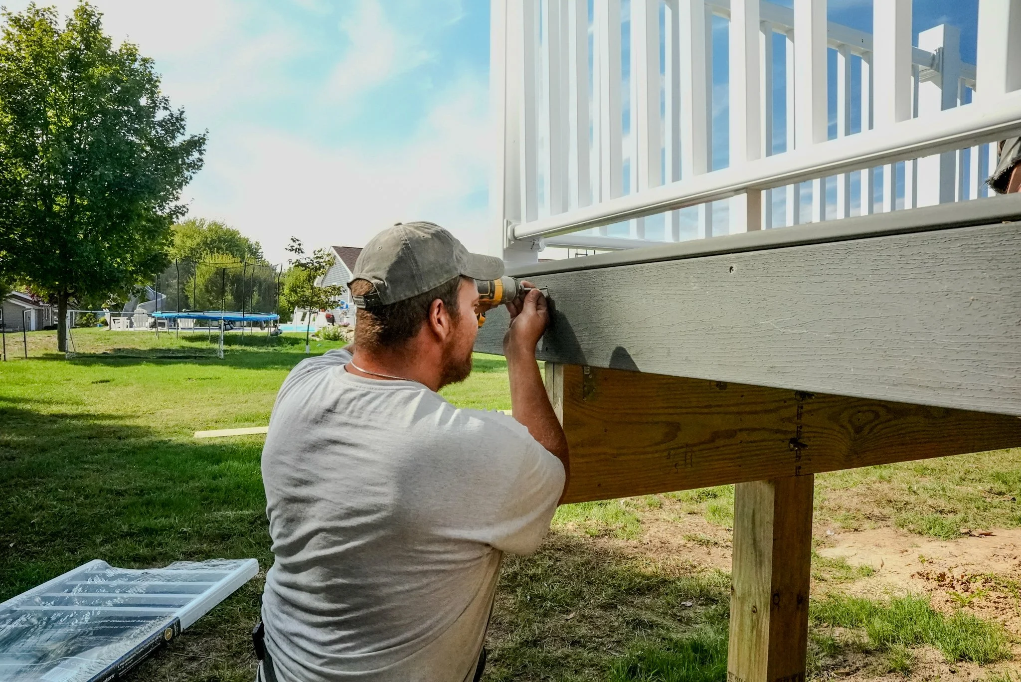 Person installing or repairing a railing on a raised deck using a drill outdoors in a backyard.