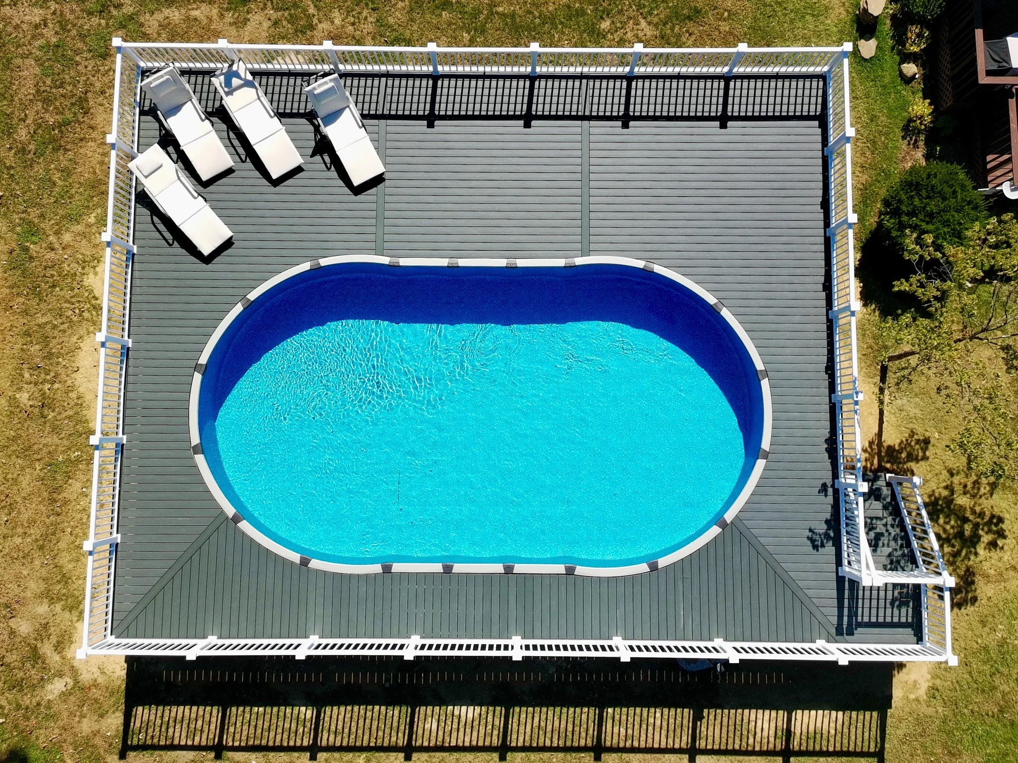 An aerial view of a house with a large screened-in backyard deck and an oval-shaped swimming pool with blue water. Four white lounge chairs are positioned on the upper left corner of the deck, and a white gate is on the lower right side of the deck.