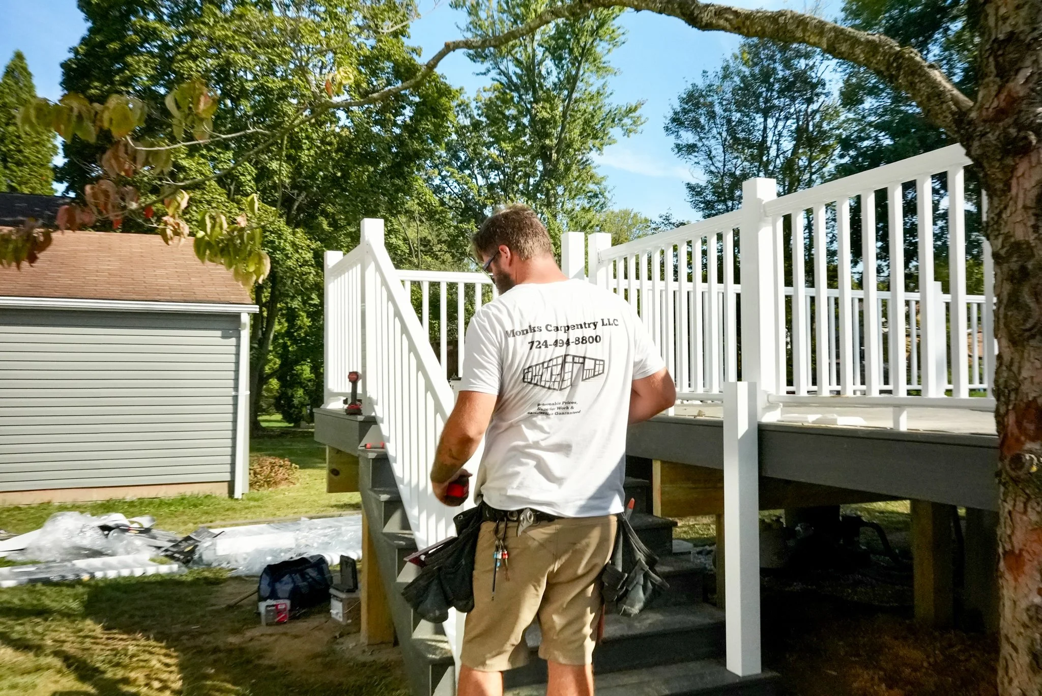 A man working on installing white railing on a wooden deck in a backyard with trees and a gray shed.