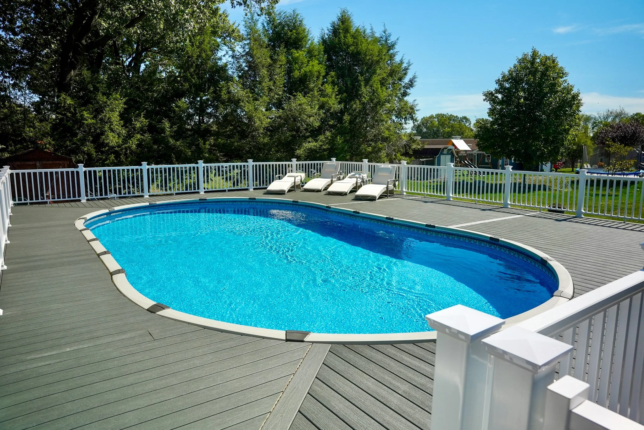An outdoor swimming pool on a deck with lounge chairs, surrounded by a white fence and green trees in the background on a sunny day.