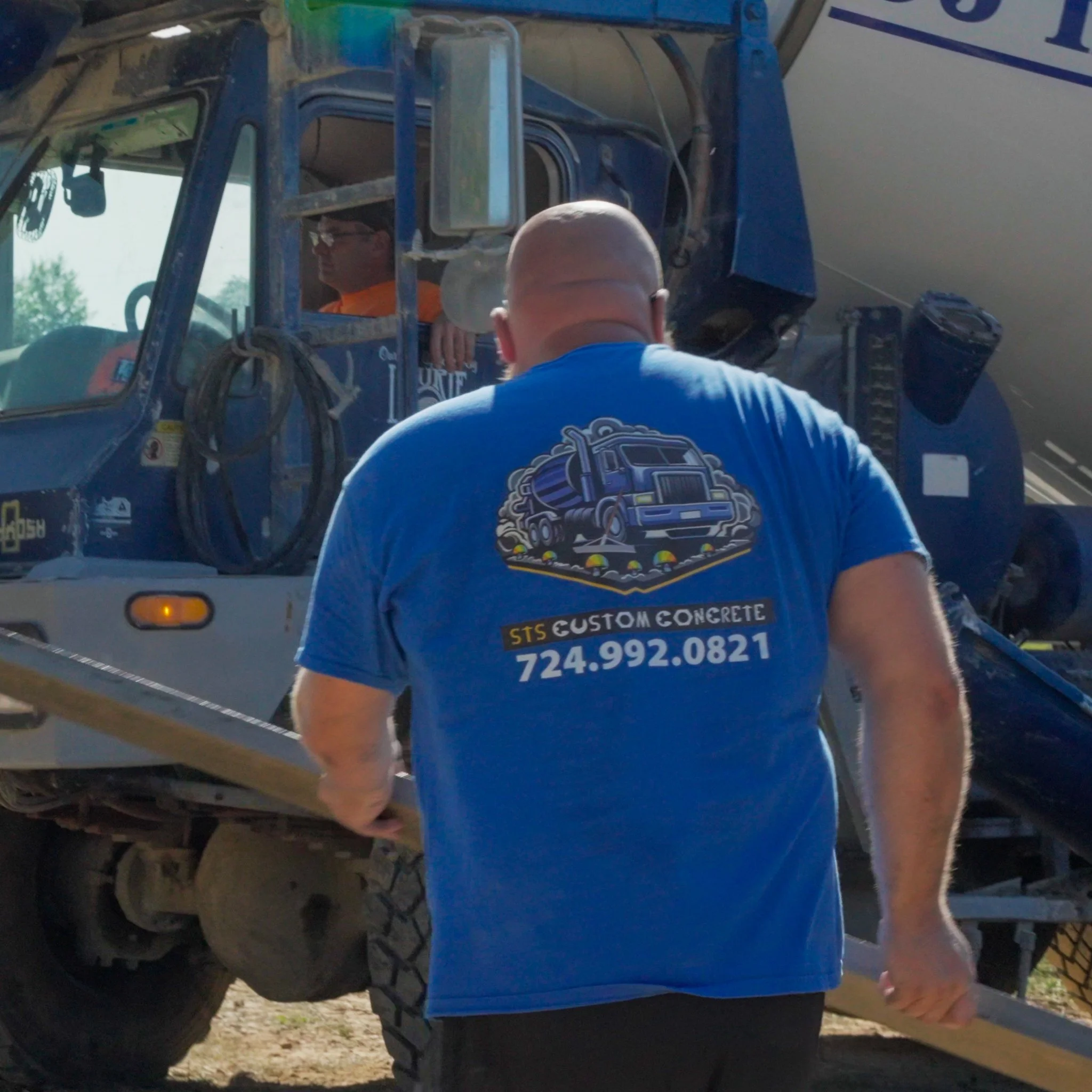 A Pittsburgh contractor seen from behind wearing a blue t-shirt with a graphic of a concrete mixer truck and the text 'STS CUSTOM CONCRETE' along with a phone number. He is approaching a large cement mixer truck at a construction site.
