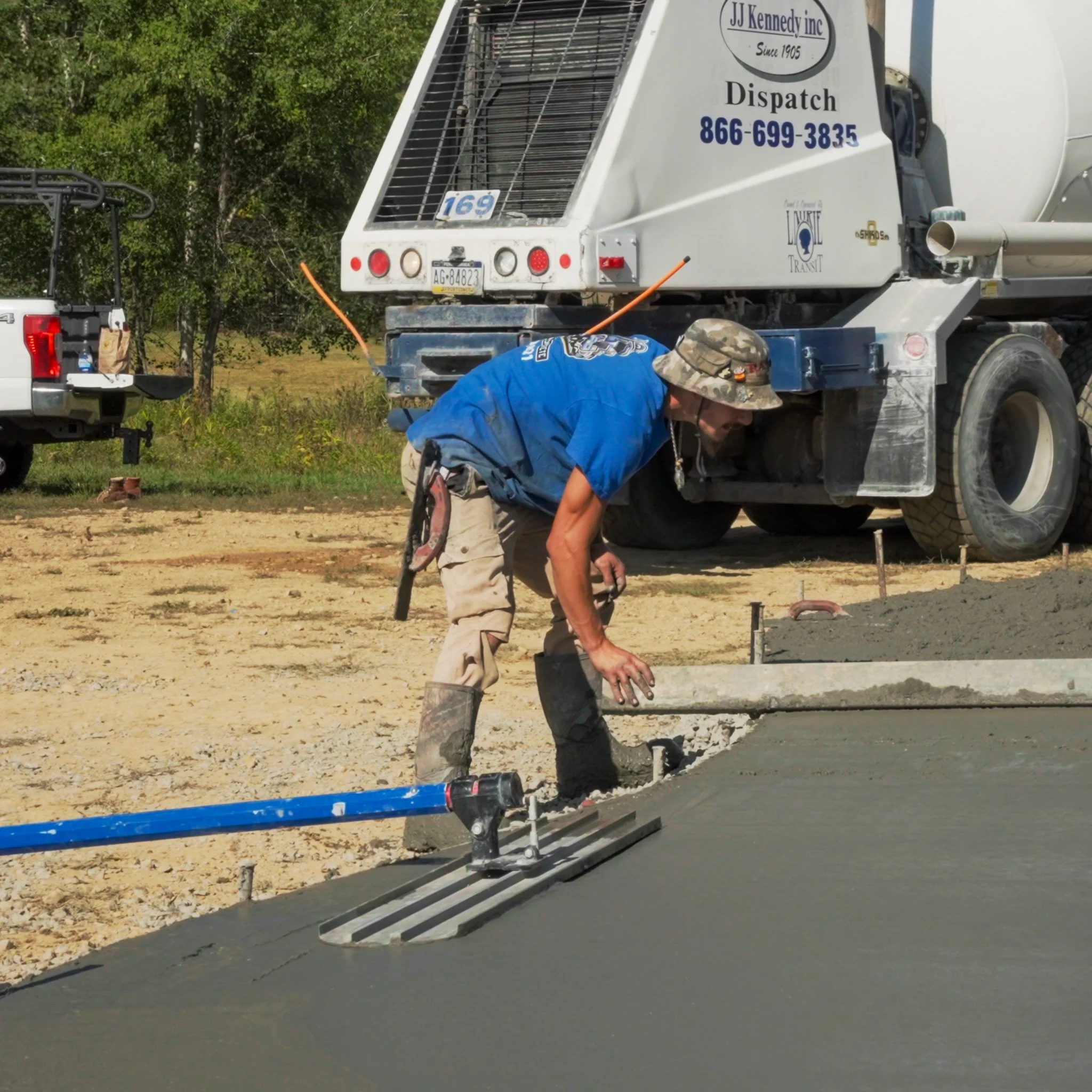 A Pittsburgh construction worker in a camouflage hat and blue shirt smoothing fresh concrete on a road, with construction equipment and parked trucks nearby.