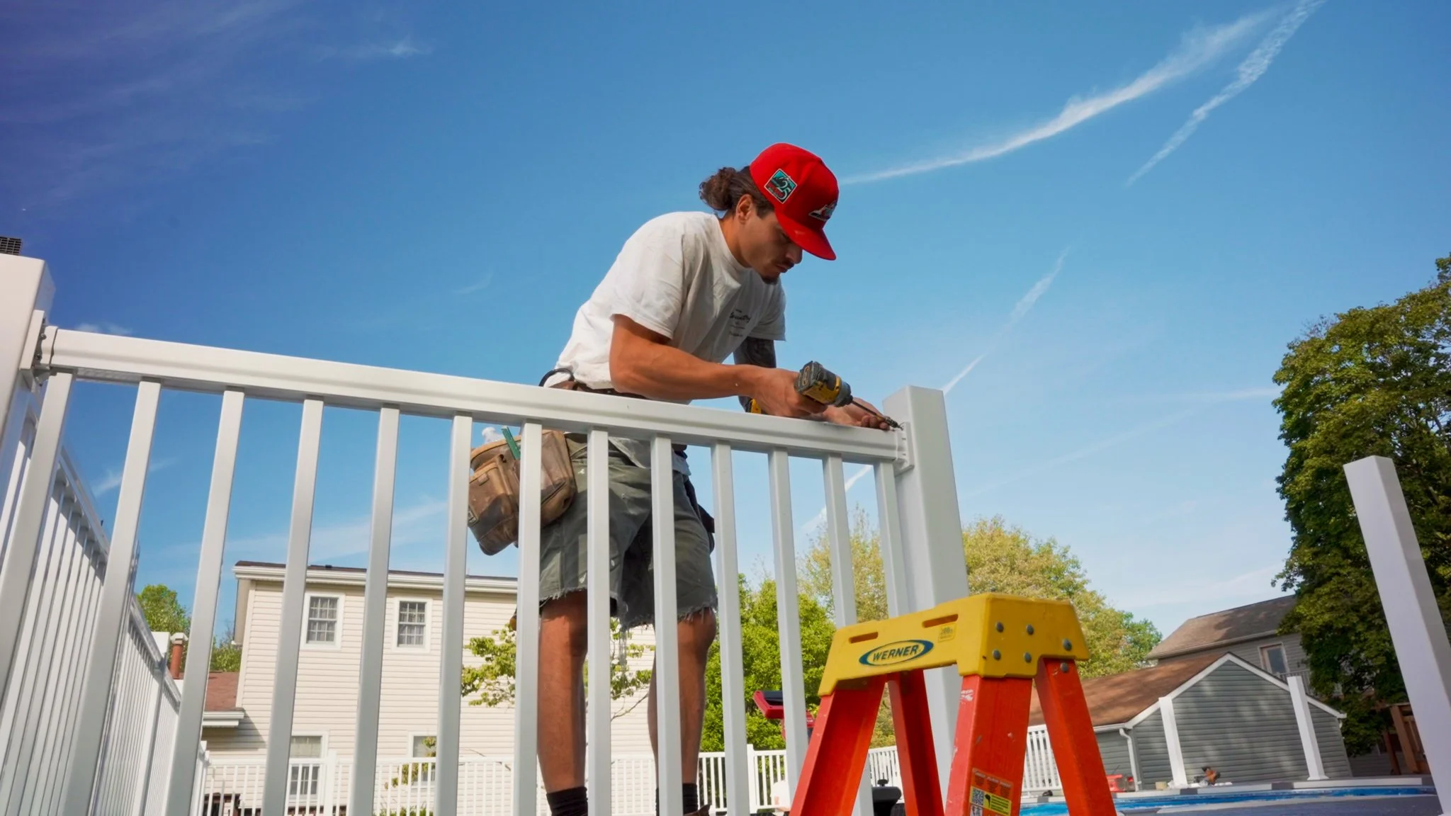A man installing or repairing a white fence using a cordless drill on a sunny day in a residential neighborhood.
