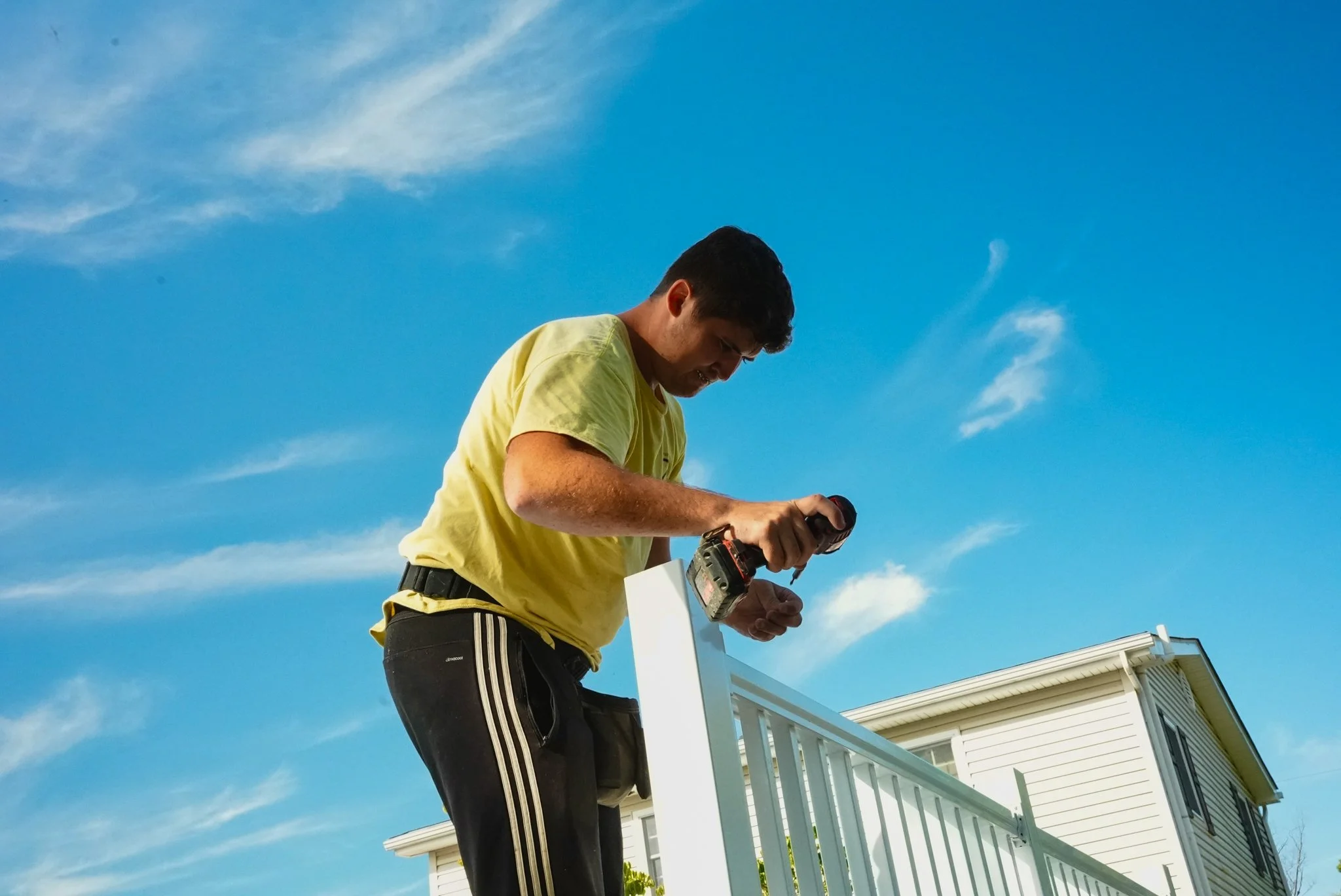 A young man in a yellow shirt and black pants working on a white fence with a power drill outdoors under a blue sky.