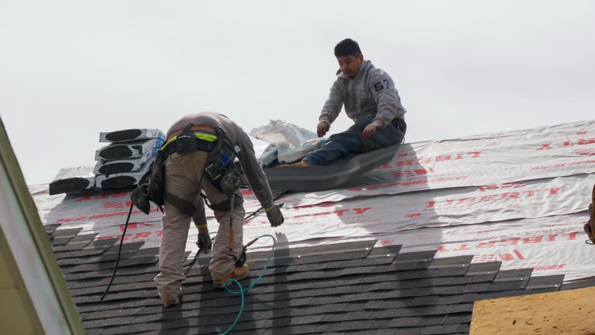 Two Pittsburgh roofers working outdoors, one with a white hoodie with a business logo and the other with a red hoodie, working on a roof repair project.