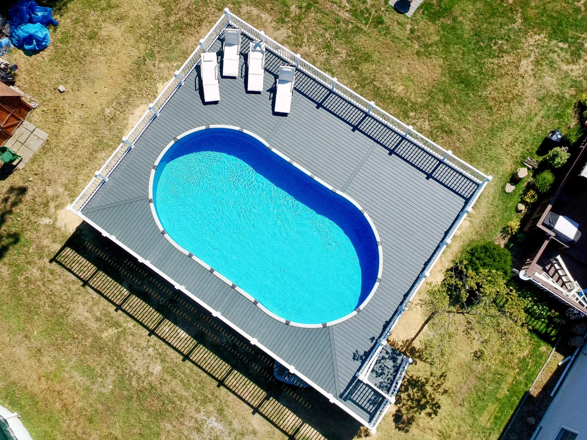 Aerial view of a backyard with a rectangular gray deck and a kidney-shaped swimming pool with clear blue water. There are five white lounge chairs on one side of the deck and lush green grass surrounding the area.