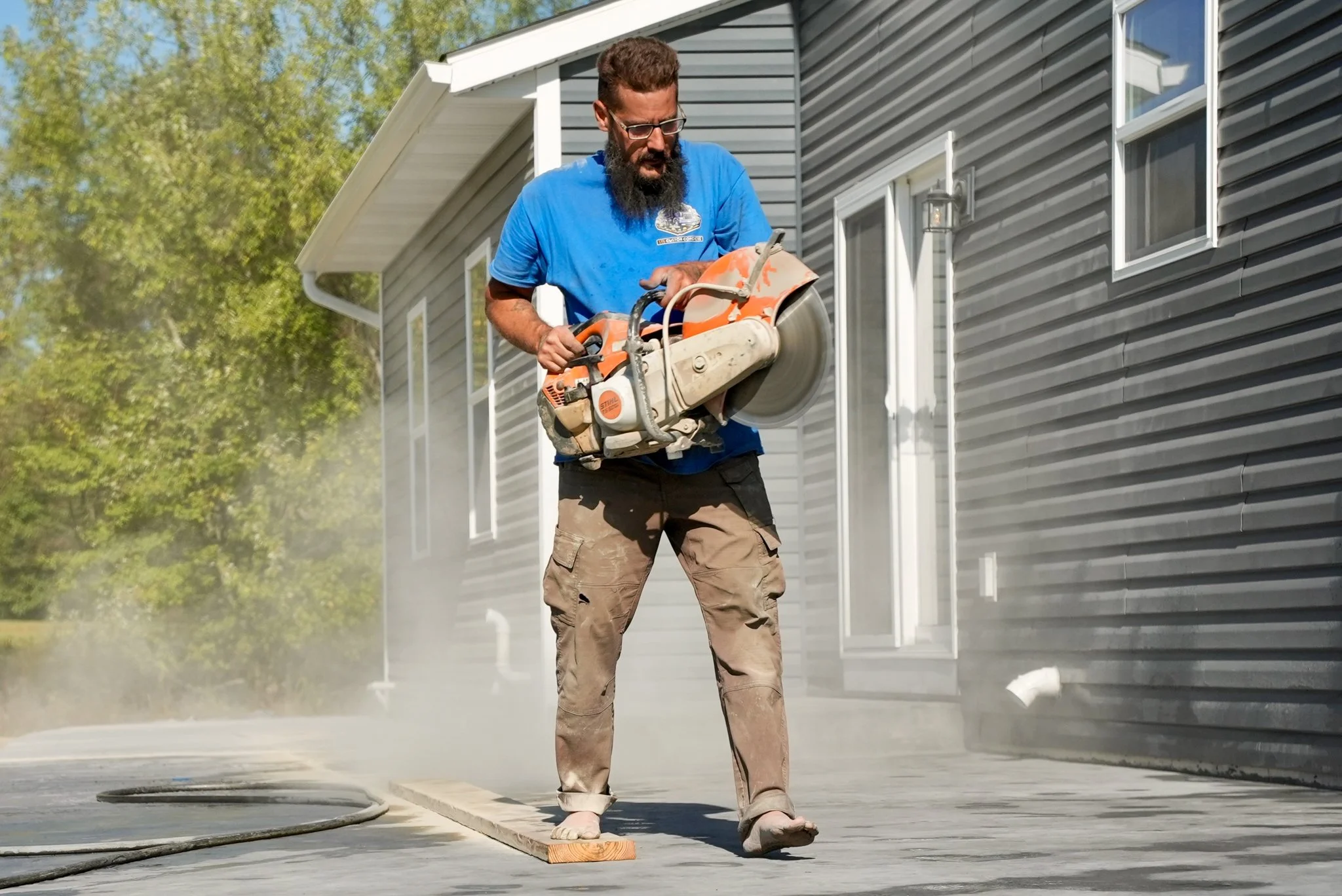 Pittsburgh contractor, wearing a blue shirt and khaki pants, using a concrete saw on an outdoor concrete surface beside a gray house, with green trees in the background.