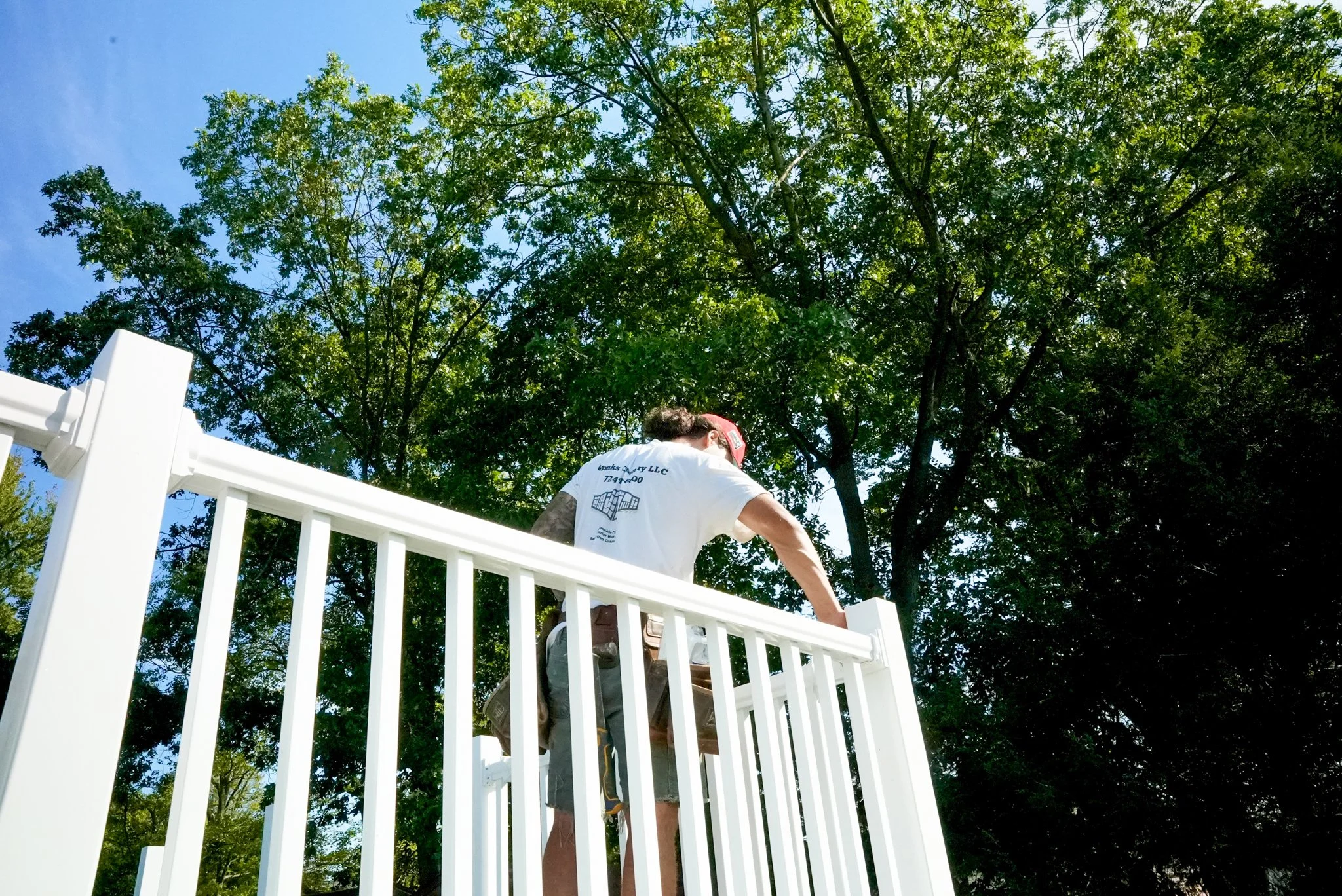A man wearing a white T-shirt and shorts working on installing or repairing a white picket fence in a backyard, with green trees and a blue sky in the background.