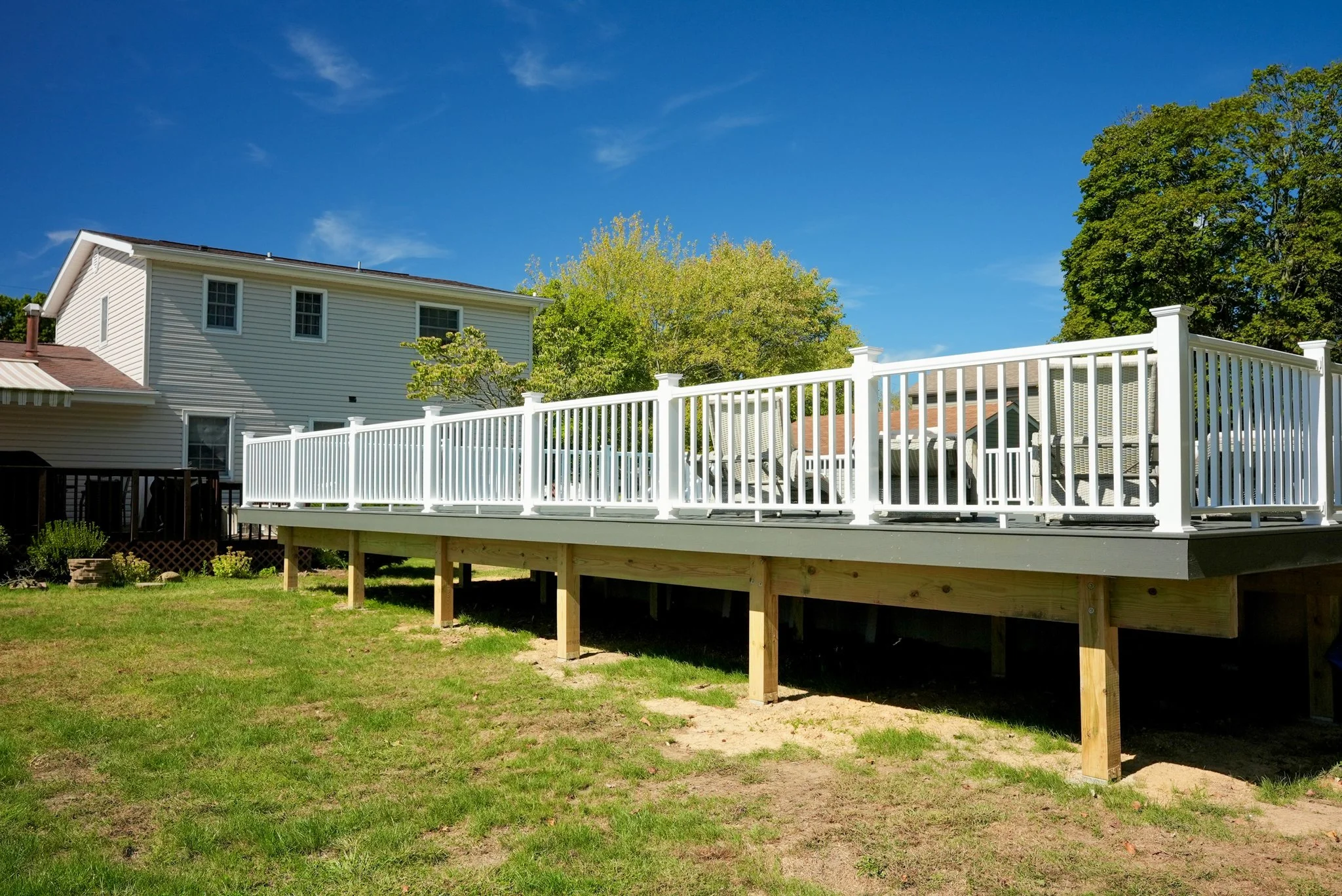 A backyard deck with white railing, supported by wooden posts, attached to a two-story white house with vinyl siding, trees in the background, and a bright blue sky.