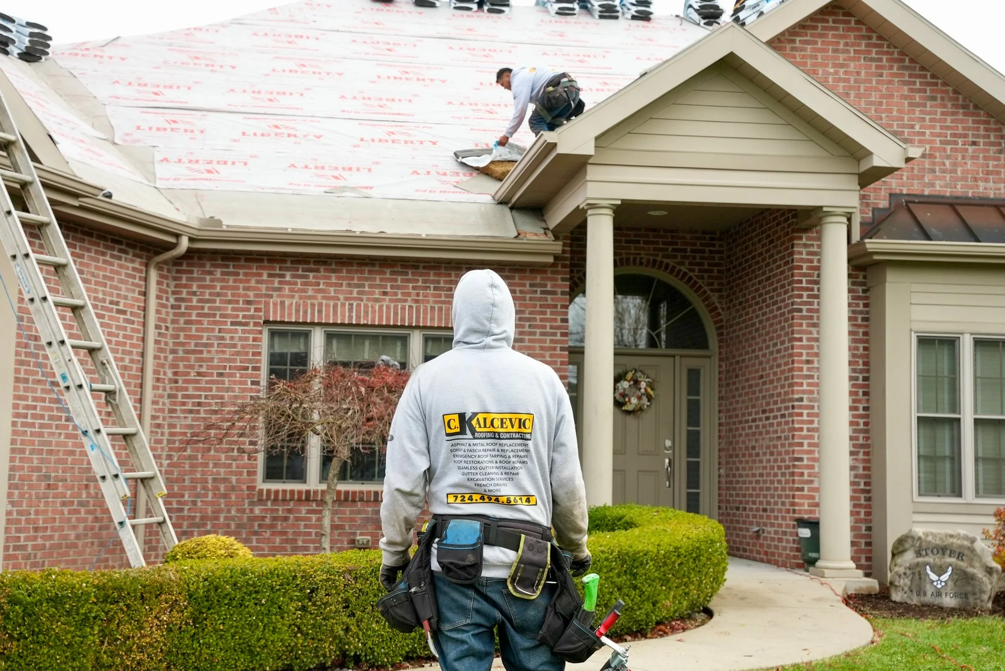 A Pittsburgh roofing contractor inspecting the roof of a brick house, with one worker on the roof and another standing on the ground looking up. The worker on the roof is kneeling and working on the shingles, while the person on the ground is wearing