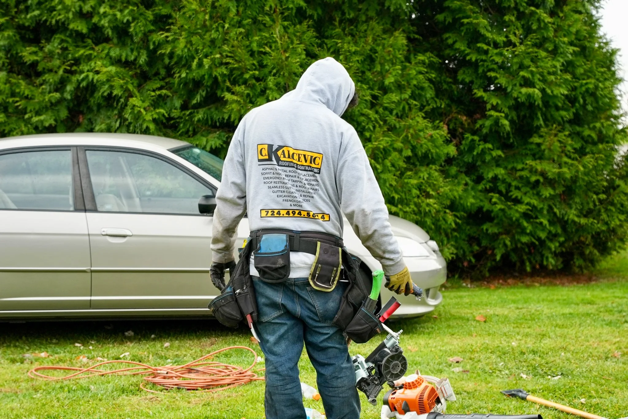 A Pittsburgh roofer dressed in a gray hoodie and jeans, with tools attached to their belt, is standing on grass near a silver car and green bushes. The worker is holding a tool and appears to be doing some work, with equipment and extension cords on 