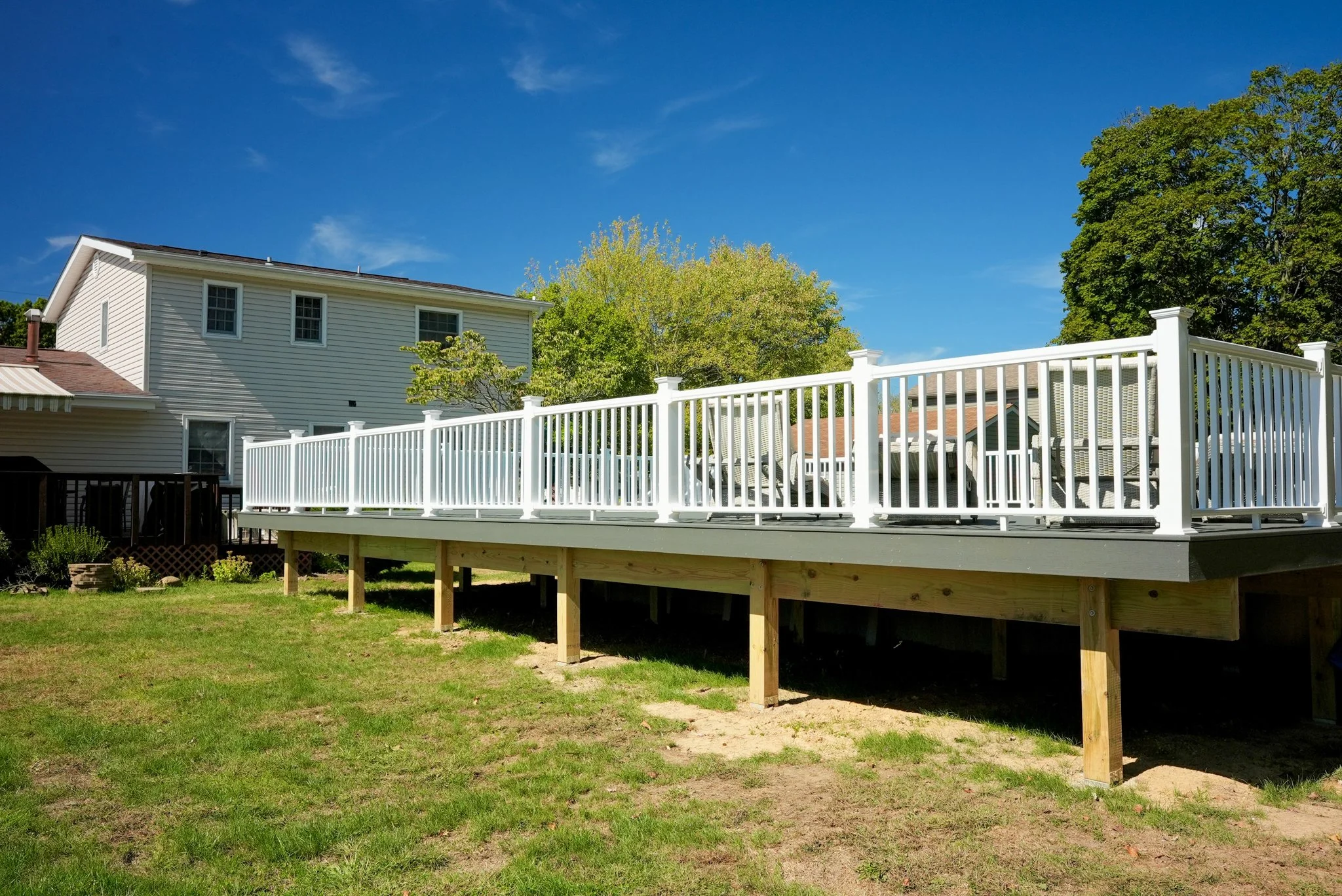 Residential backyard with a newly built elevated deck supported by wooden posts, featuring white railings, set against a white house with an awning, a green lawn, and trees under a blue sky.