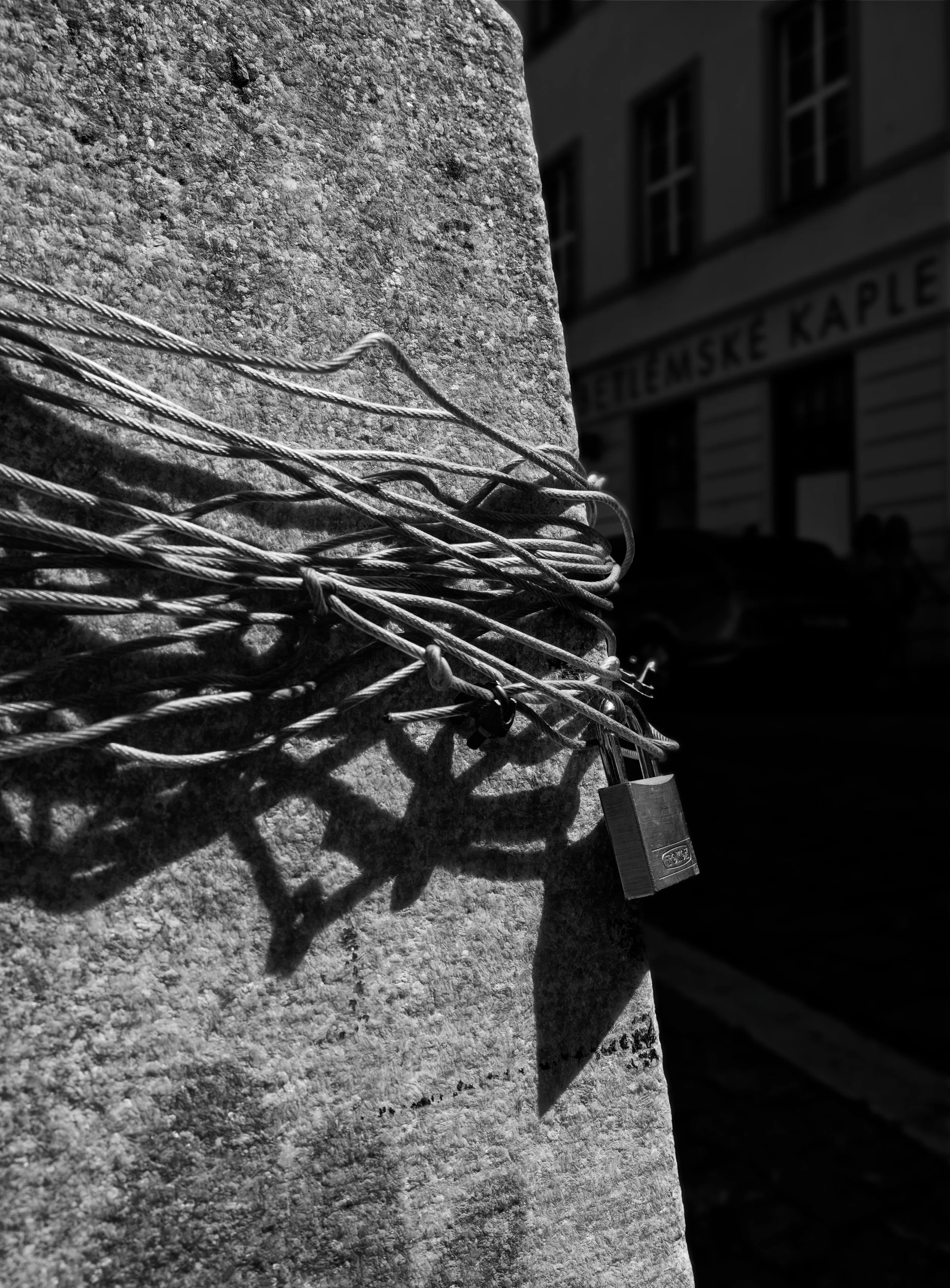 A tangle of steel cables and a padlock wrapped around a stone pillar in Prague, casting sharp shadows in strong sunlight.