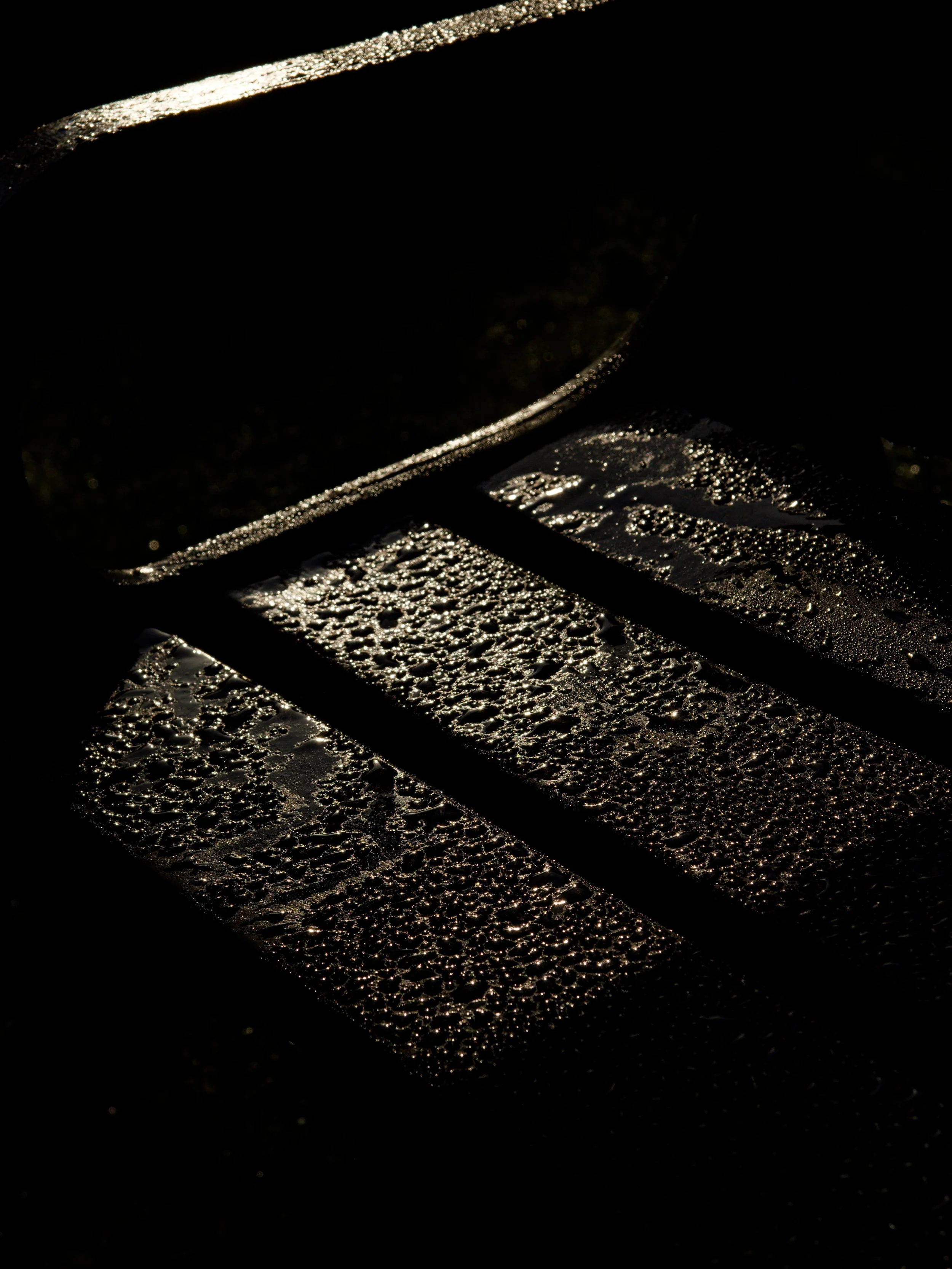 Sunlight catches raindrops on a dark bench, creating bright textured highlights and deep geometric shadows.