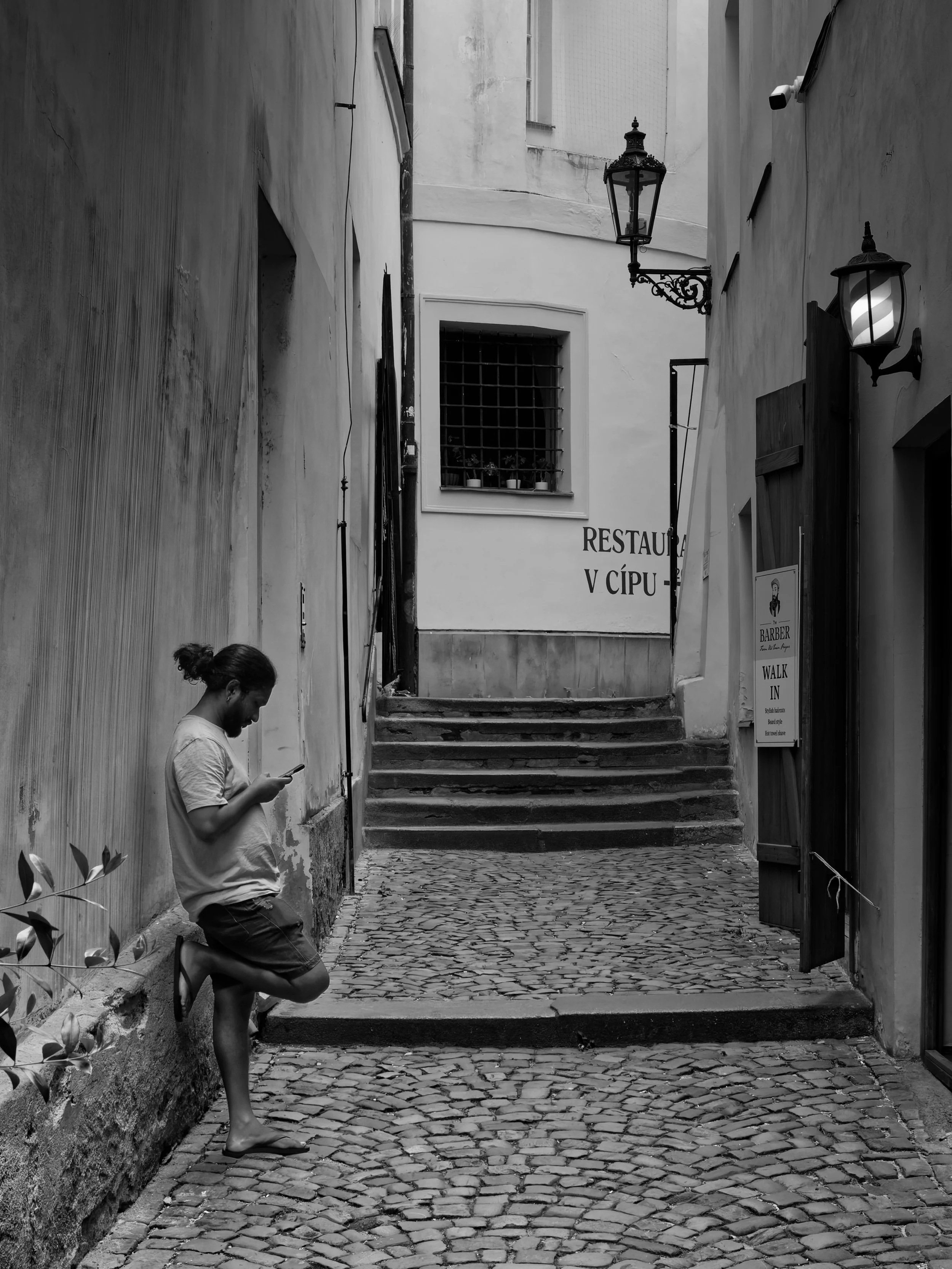 A person pauses to look at their phone in a narrow cobbled passage in Prague, framed by worn walls, steps and old street lamps.