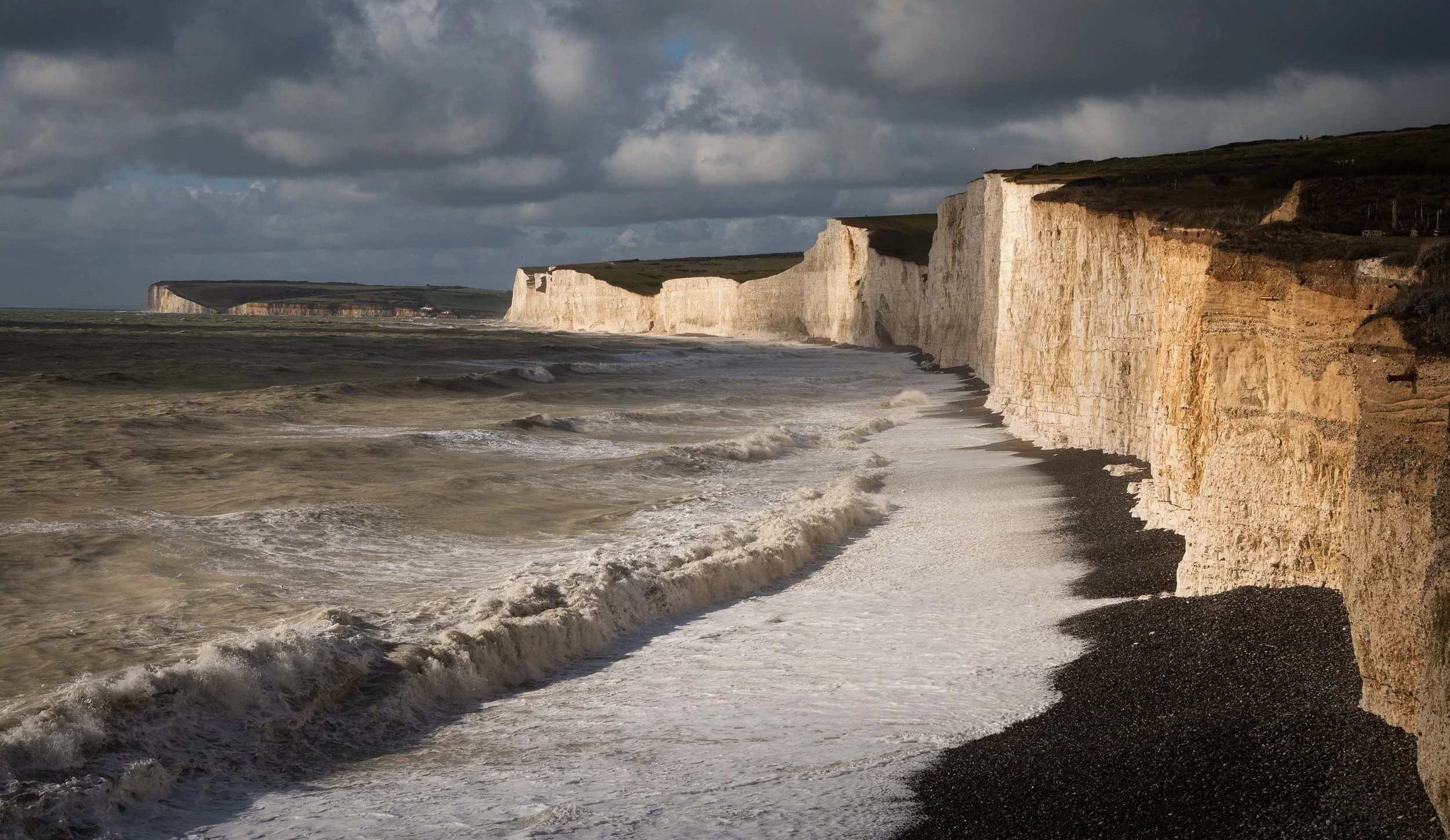 Cliffs along a rocky shoreline with waves crashing against the beach and dark clouds overhead.