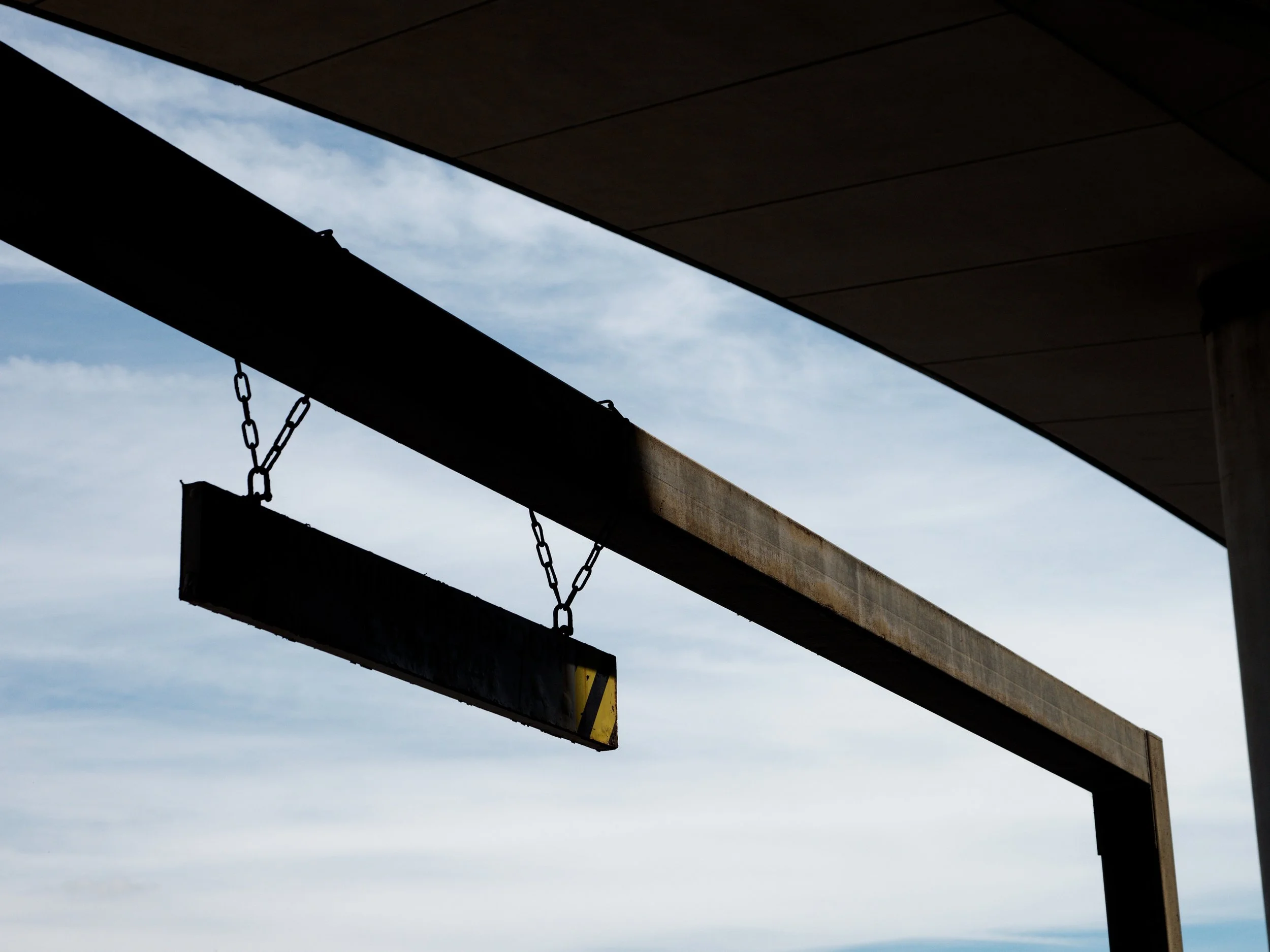 A suspended height-restriction bar hangs from chains beneath the concrete structure of a car park at Brighton Marina, silhouetted against a pale blue sky.