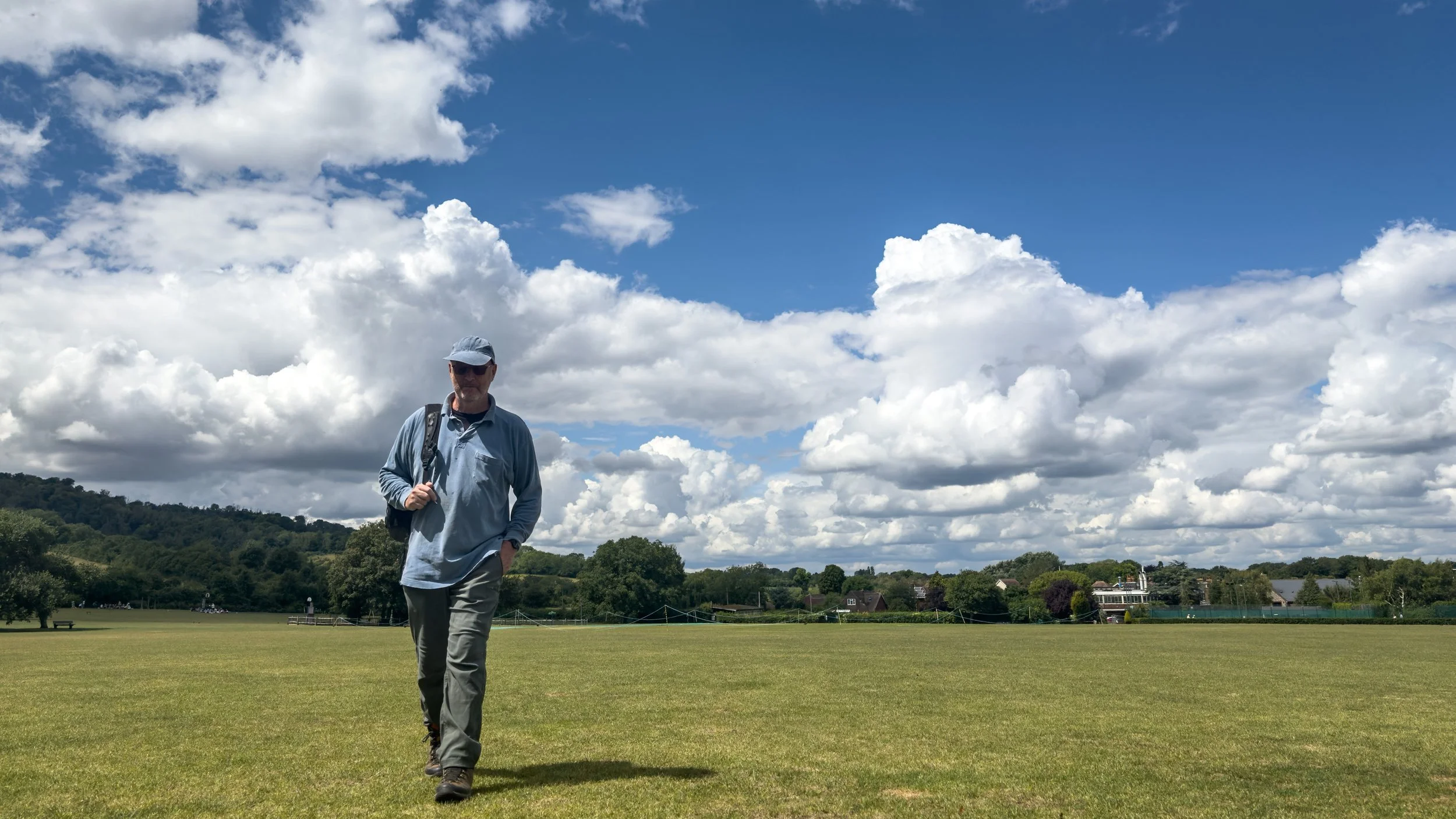 Man wearing sunglasses, a blue hat, and a light blue jacket walking on a grassy field with trees and houses in the background under a partly cloudy sky.