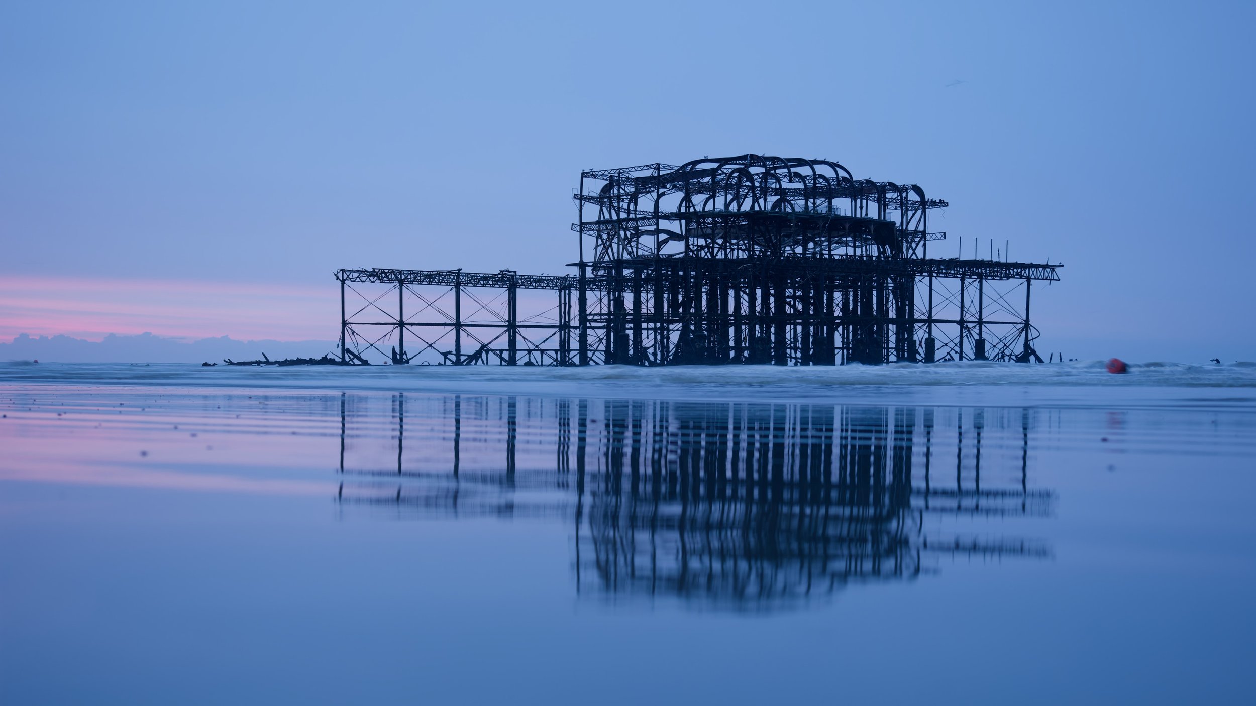The skeletal remains of Brighton West Pier stand reflected in wet sand during blue hour, with gentle waves and a calm morning sea.