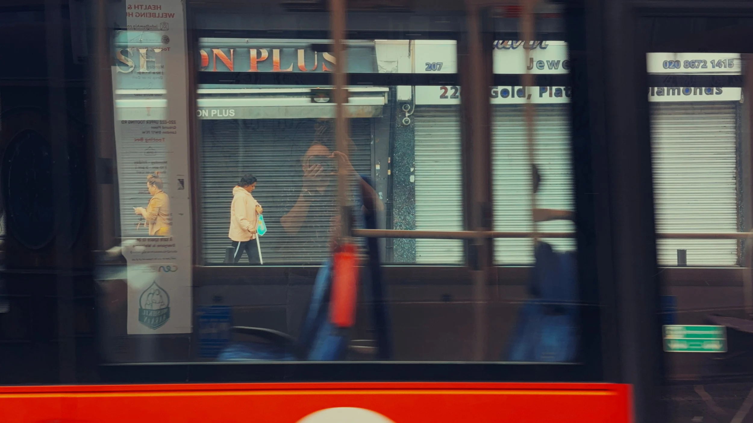 Pedestrians, reflections and the photographer briefly align through the windows of a passing London bus on Tooting High Street.