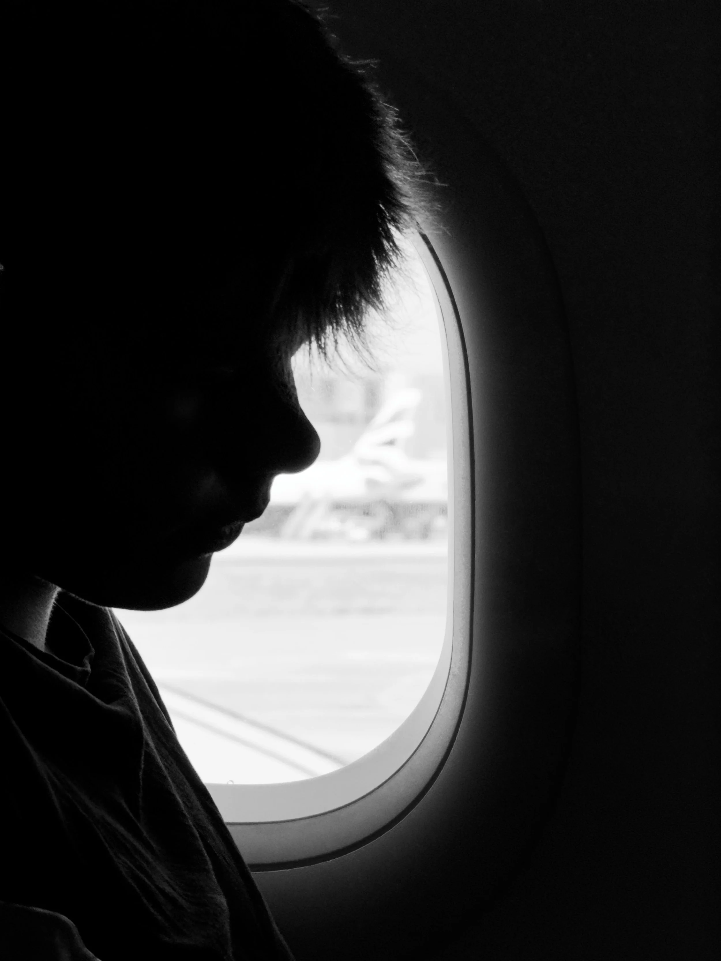 A young passenger sits in silhouette beside an aircraft window, their profile softly outlined by the bright light outside.