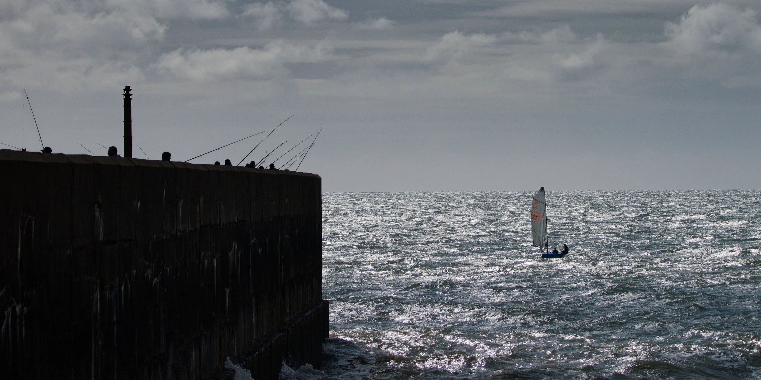 A small sailing dinghy crosses rough, sunlit water beside the harbour wall, dwarfed by the structure and the fishermen above.