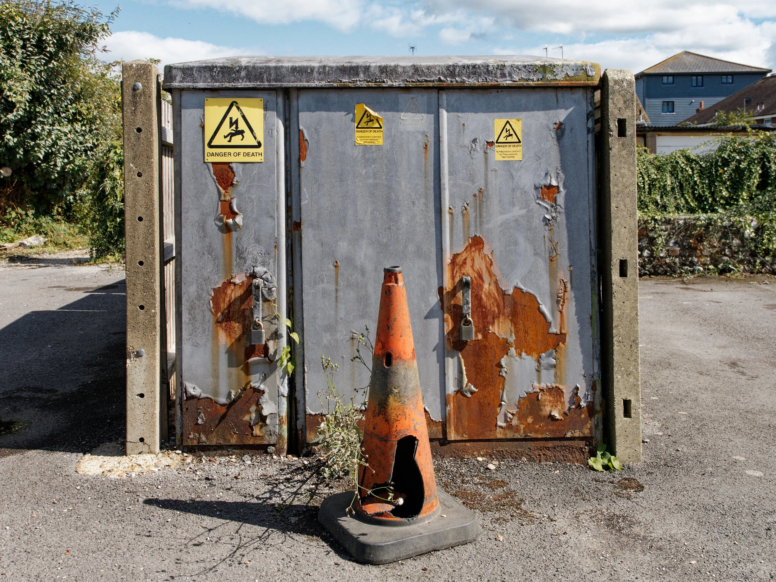 A badly damaged orange traffic cone stands in front of a rusting grey electrical cabinet displaying three yellow Danger of Death warning signs.