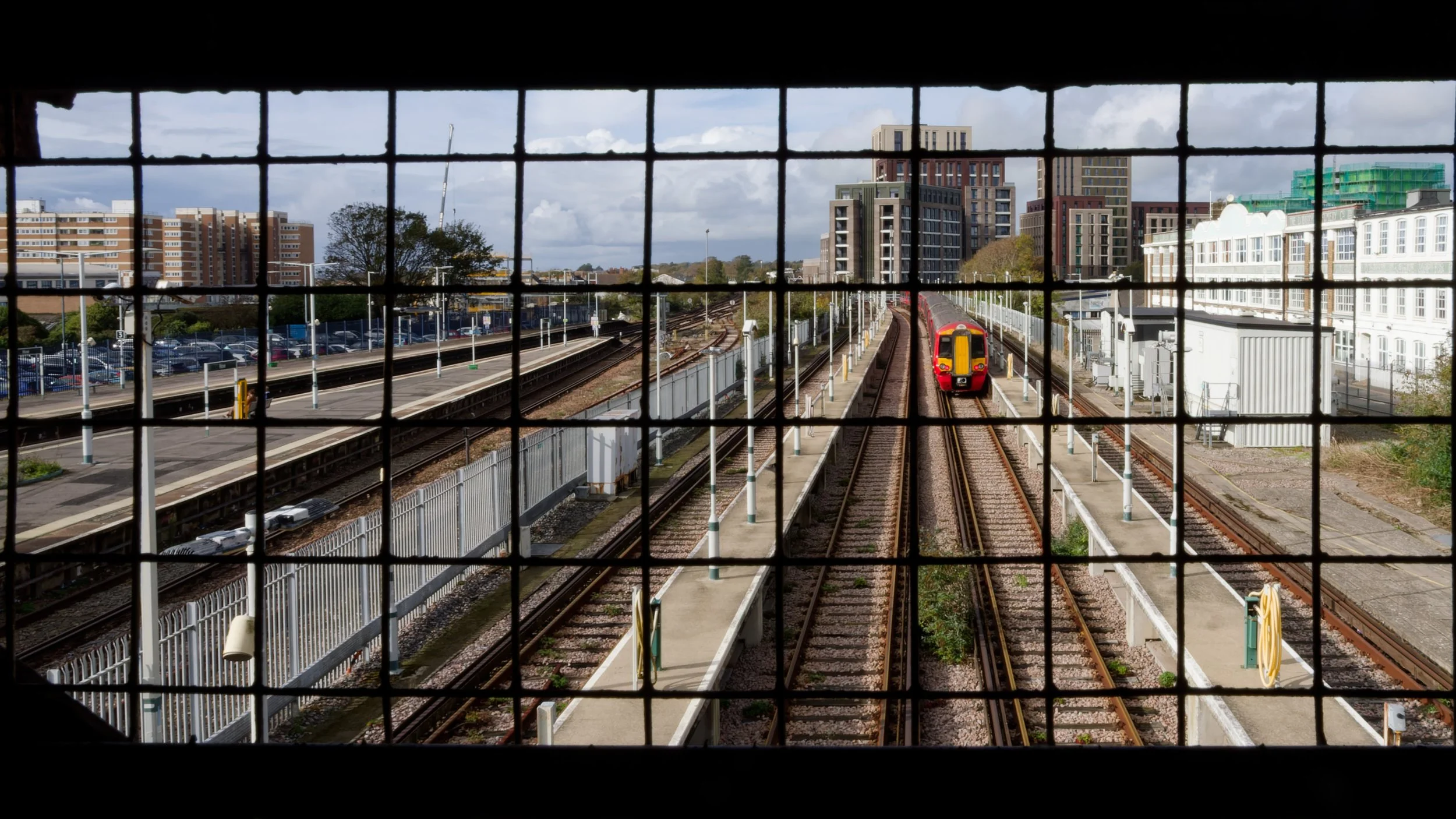 A red passenger train approaches along railway tracks, seen through a protective metal grid in an eye-level opening on a pedestrian bridge.