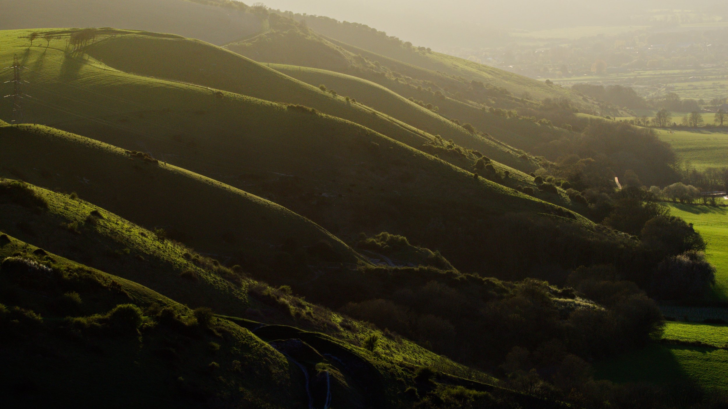 Rolling green hills in low golden light, with long shadows falling across the folds of the landscape and haze softening the distant Weald.