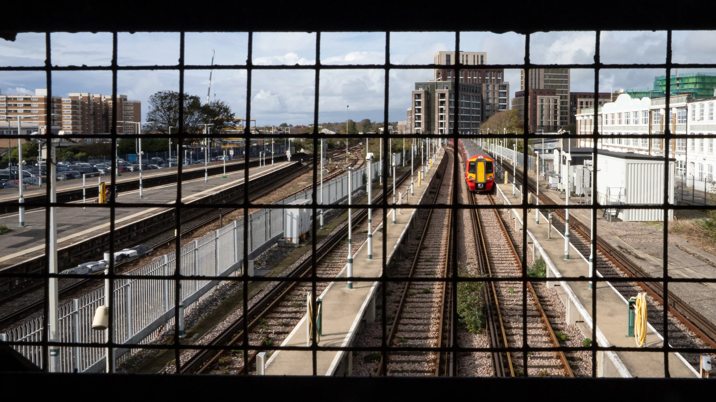 View of train tracks and a red subway train approaching on the right, seen through a metal grid or fence, with multiple buildings and a parking lot in the background.