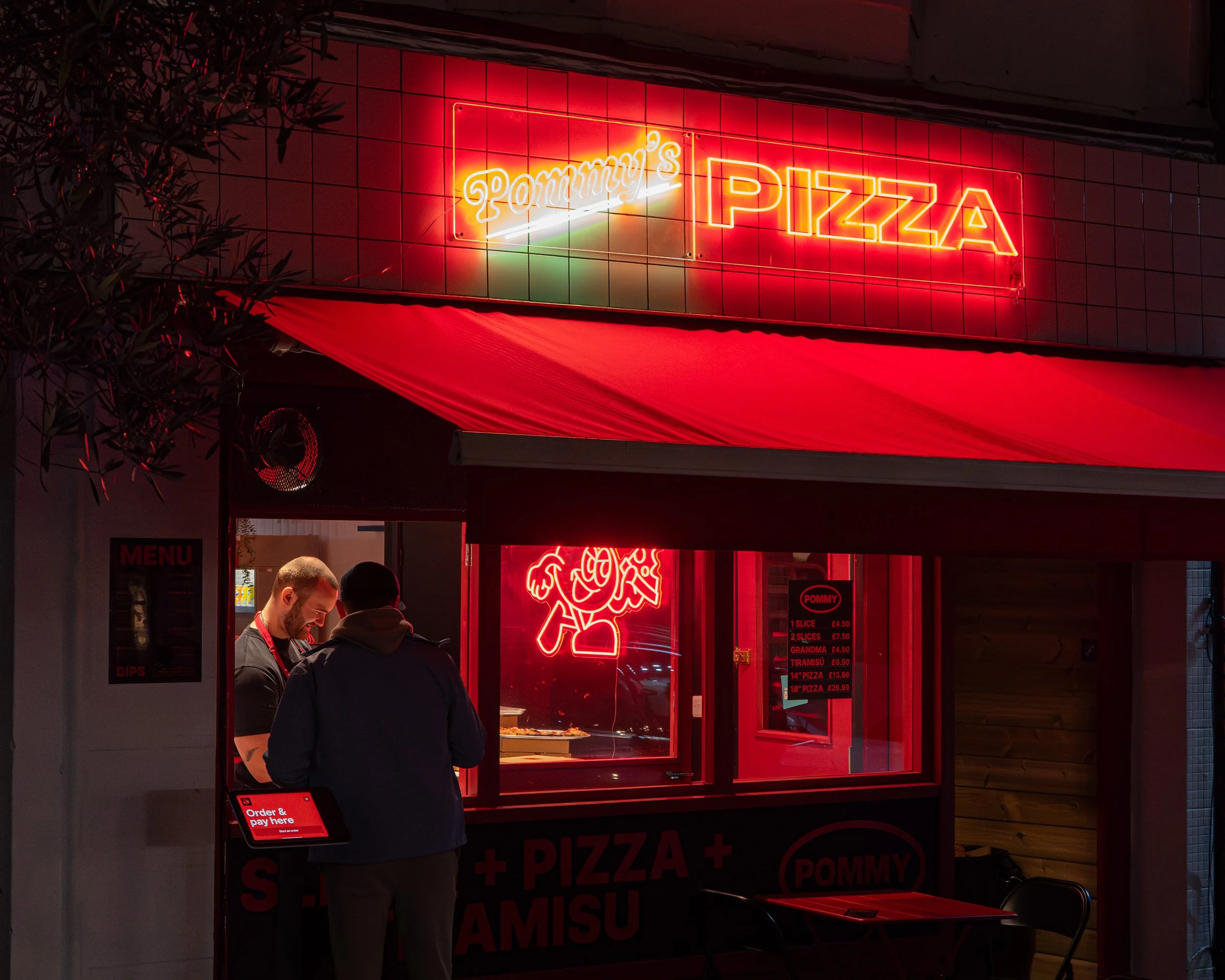 A neon sign reading 'Tommy's PIZZA' in red and white, above a pizza shop with a red awning. Two people are standing at the counter inside, one of whom is wearing a dark jacket. The shop interior is lit with red lighting, and a menu is visible on the wall.