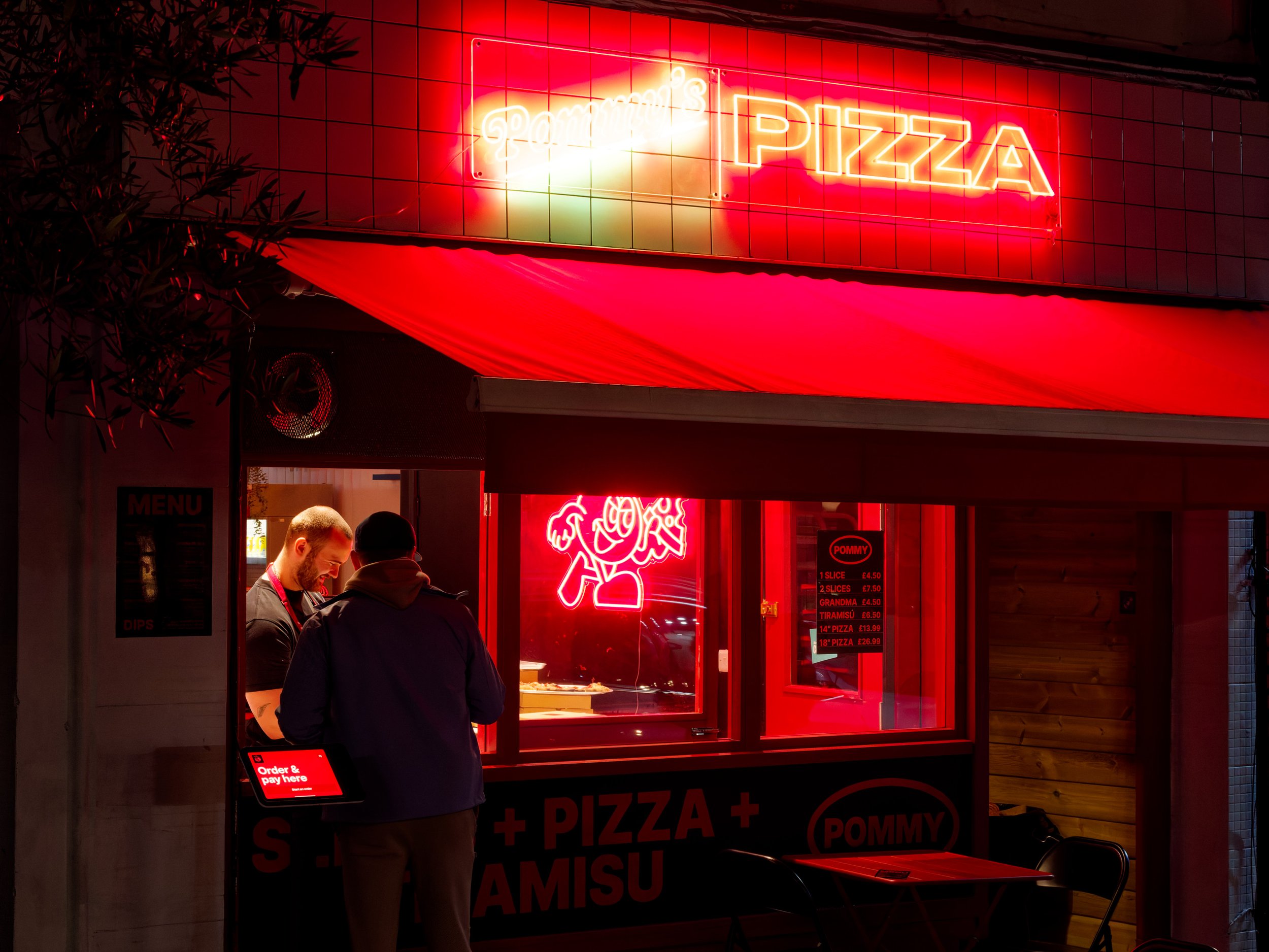 A customer orders pizza from a brightly lit takeaway beneath vivid red neon signs on an evening street.