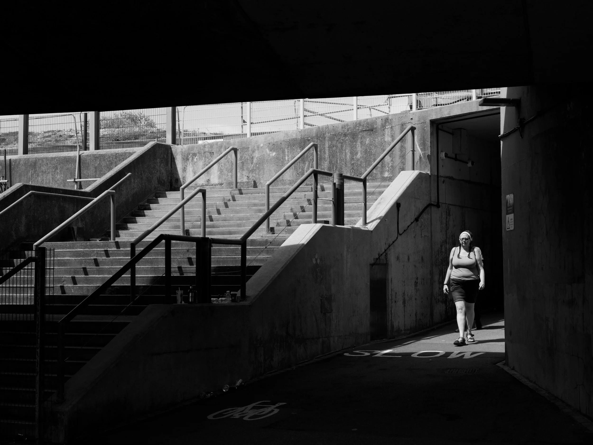 A woman walks through a sunlit pedestrian underpass at Brighton Marina, with concrete stairs and strong geometric shadows leading towards Black Rock and the seafront.