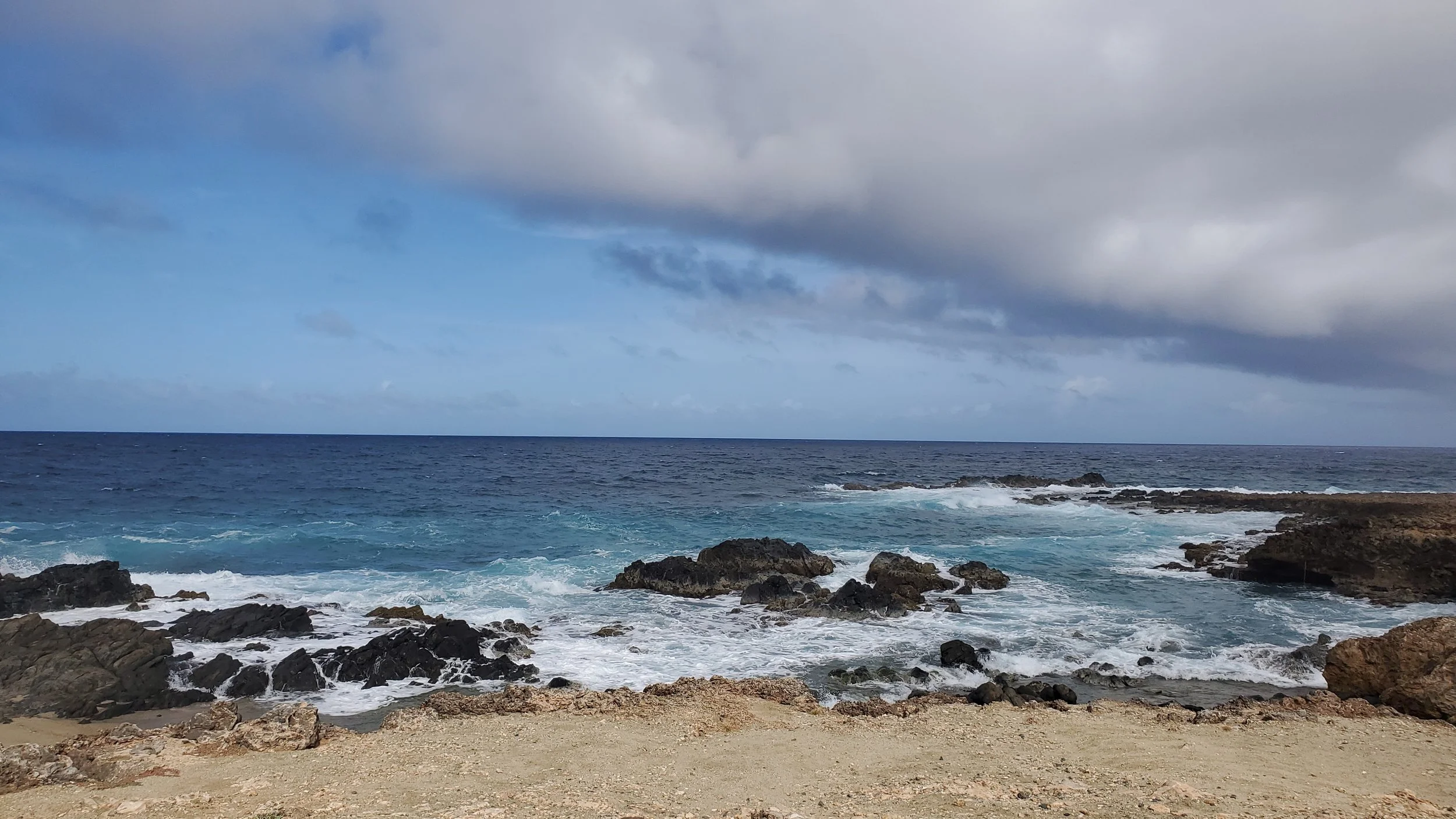 A view of the ocean with waves crashing against rocks along a sandy shore, under a cloudy sky.