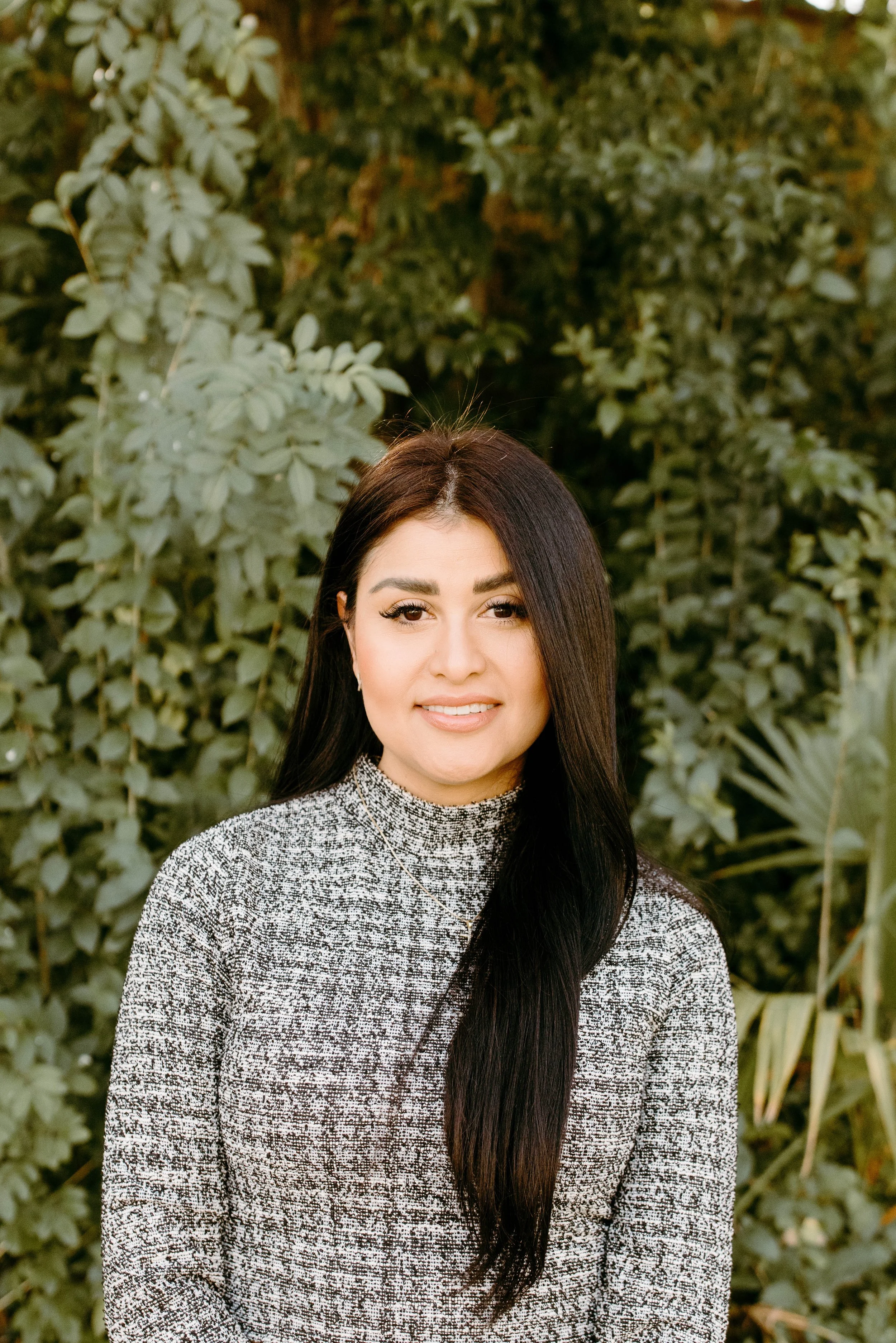 A woman with long dark hair and makeup, wearing a high-neck patterned sweater, standing outdoors with green foliage in the background.
