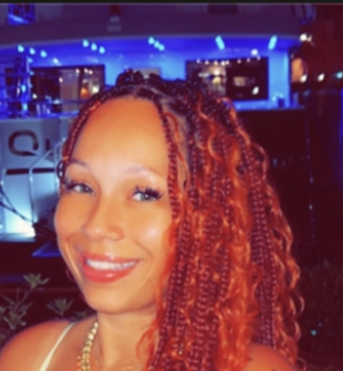 A smiling woman with curly red hair and makeup, indoors with blue lighting and television screens in the background.