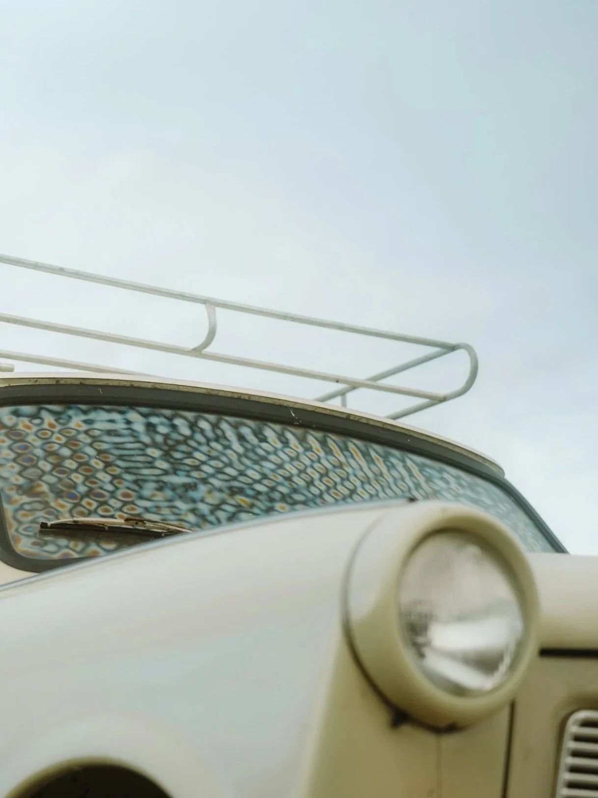 Close-up of the dashboard of a vintage car, showing a beige instrument panel and a headlight control dial. The car's windshield is partially visible, with a colorful mosaic pattern on the top edge and a metal roof rack against a pale sky background.