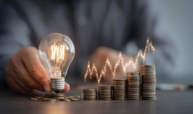A person holding a light bulb beside stacks of coins with a rising financial graph overlay.